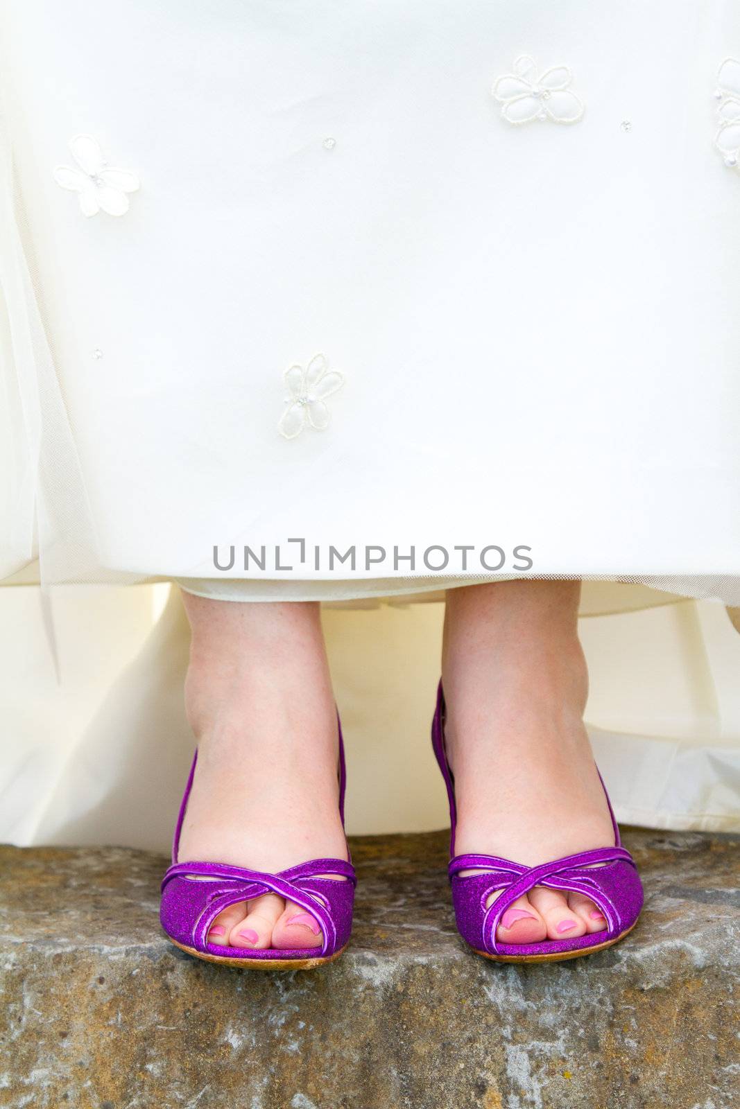 A beautiful bride in a white wedding dress stands outdoors with her pretty purple wedding shoes on her feet.