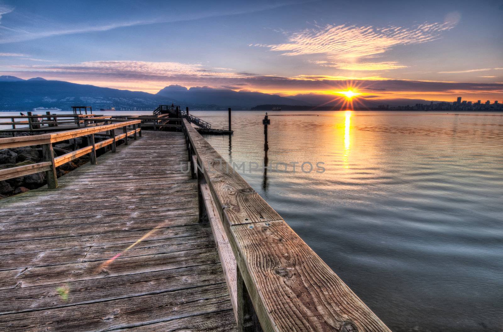 Pier moving out to the water while sun star rises over the mountain and Vancouver skyline can be seen to the right of the photo.