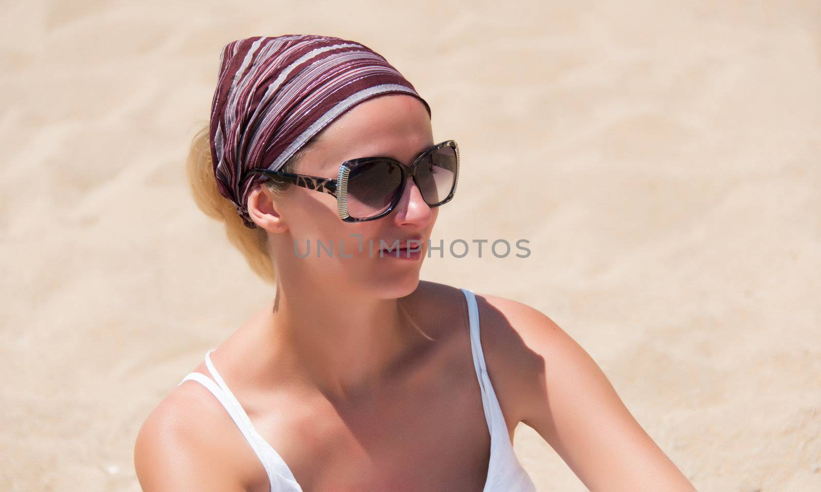 Smiling young woman in sunglasses, bandana, white dress sitting on sandy beach