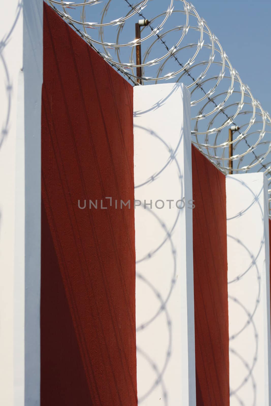 Razor wire and barbed wire on top of a red wall with white pillars, with the wire casting interesting shadows on the pillars.
