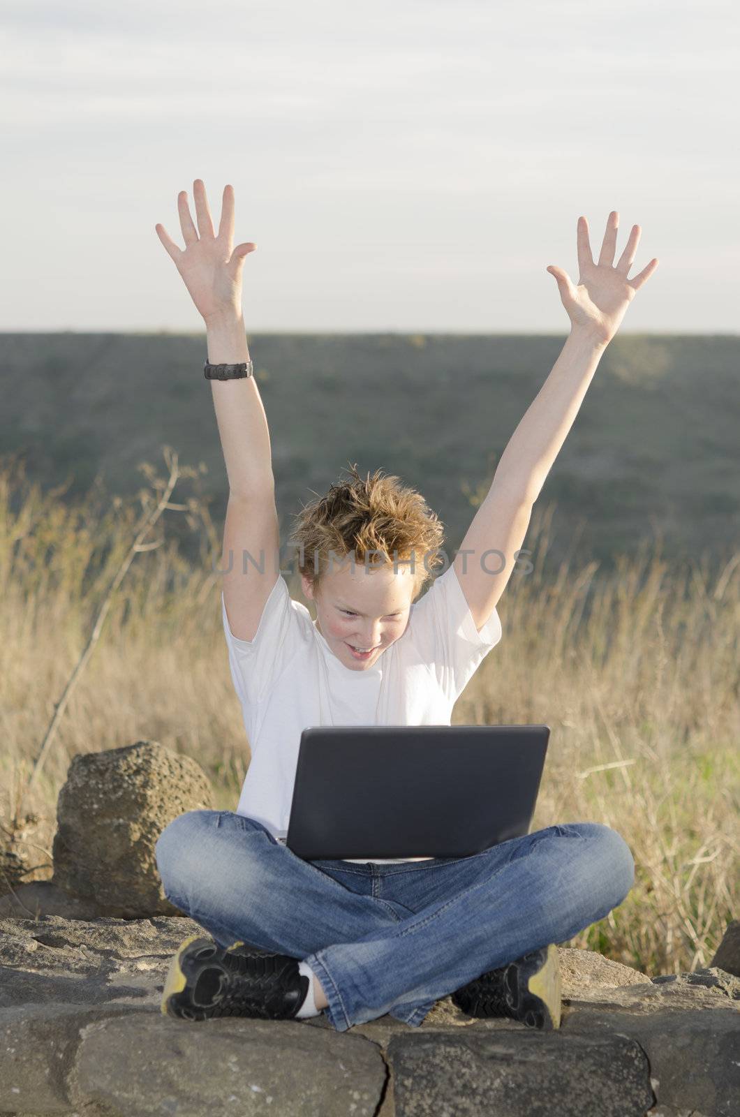 Shaggy teen with laptop joyously throws his hands up in nature