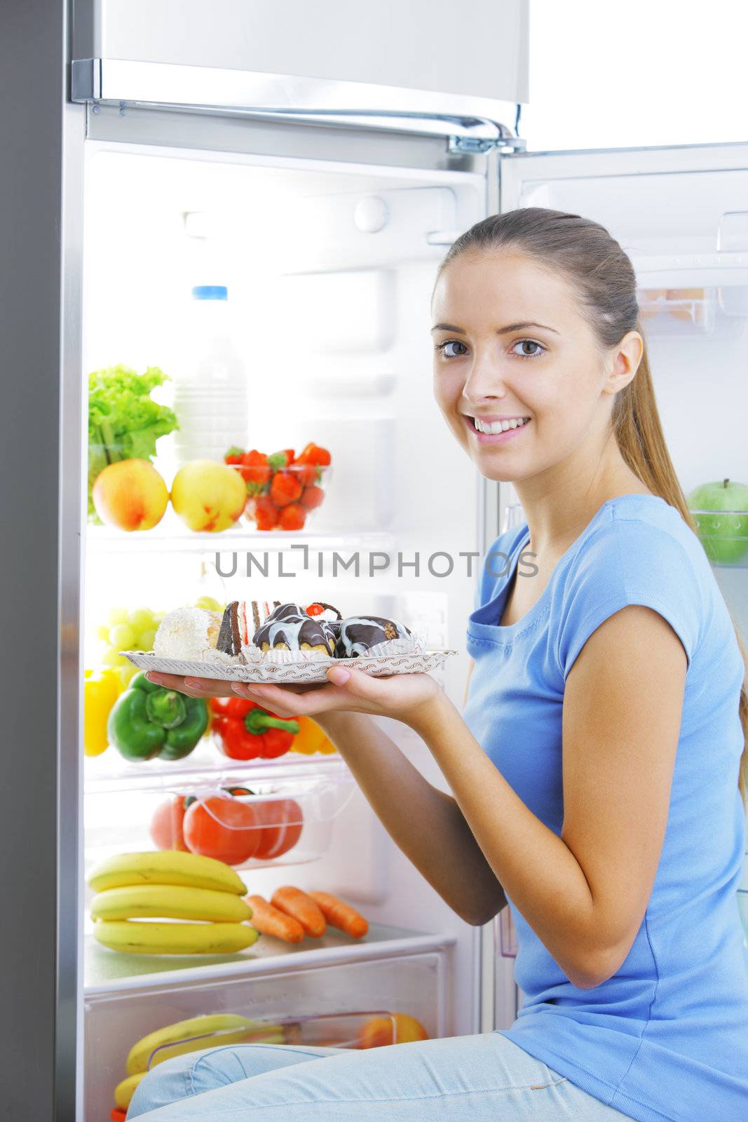 Beautiful young woman sitting near refrigerator  and choosing between healthy fruits and tasty cakes