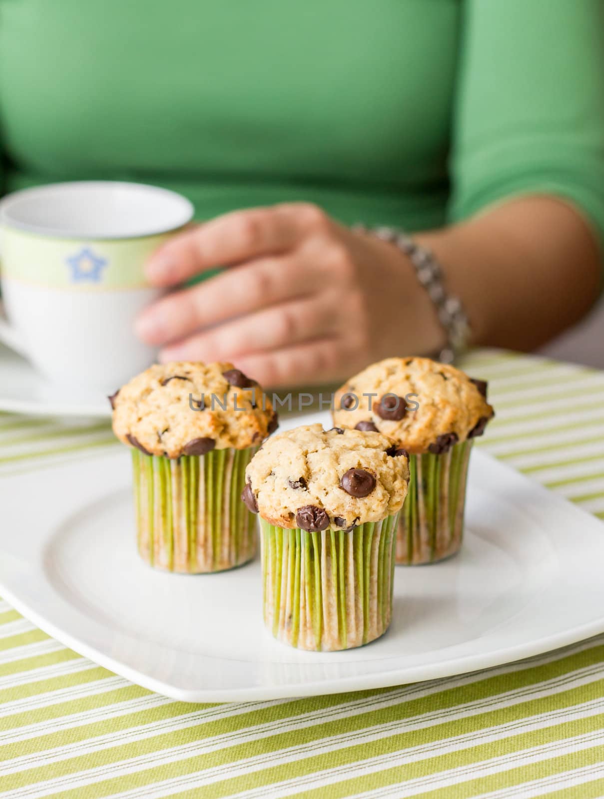 Nice girl with a cup and delicious chocolate chip muffin at breakfast in green striped tablecloth