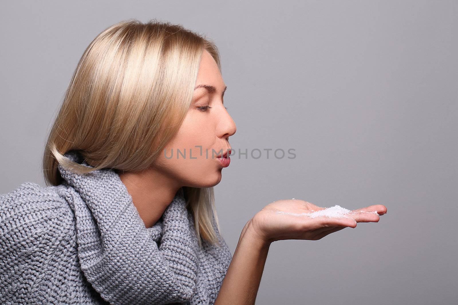 Attractive blonde blowing on snow in her hands over grey background