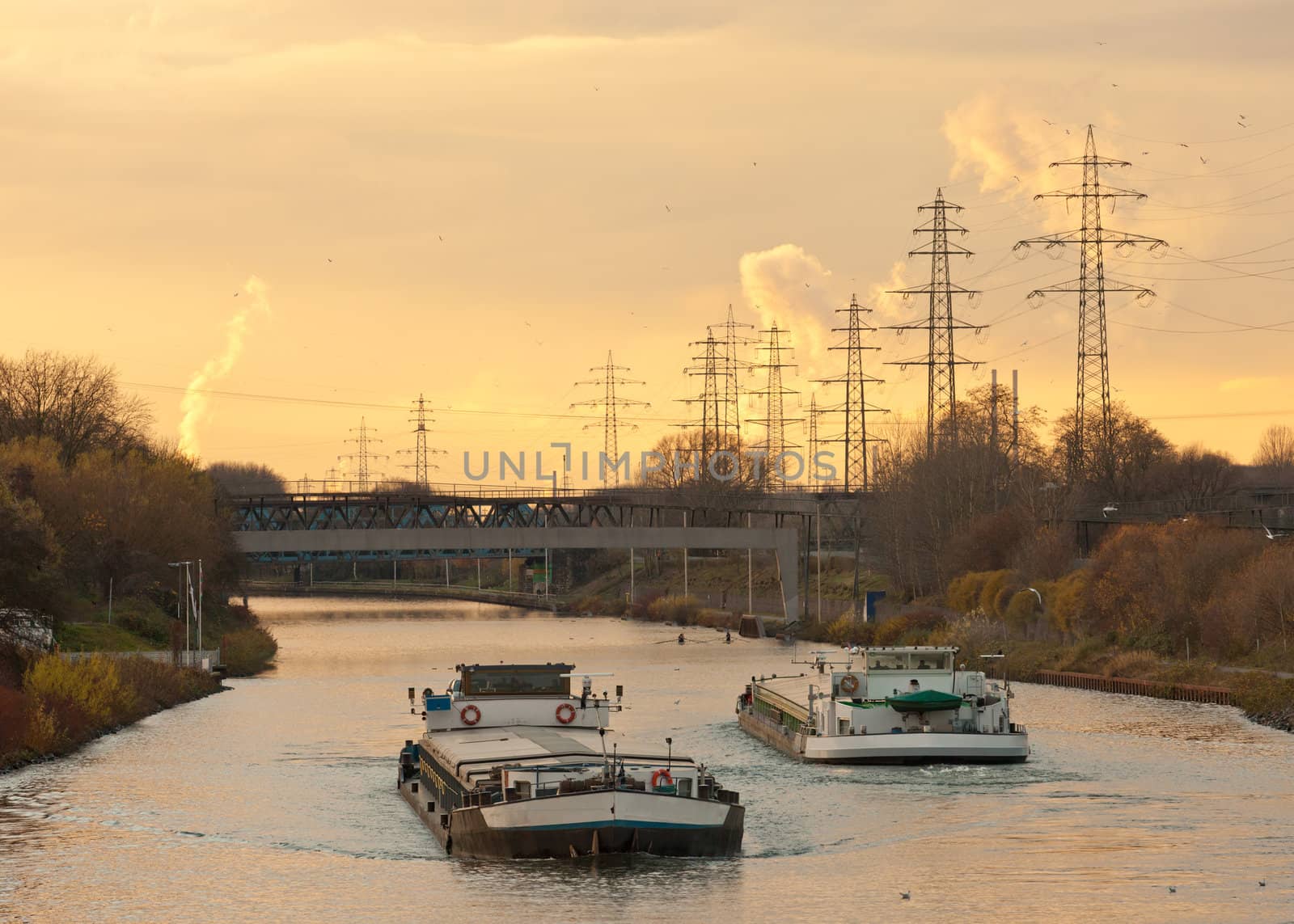 Inland waterway vessels on a busy channel in industrial area near a bridge under the warm glow of an evening sunset