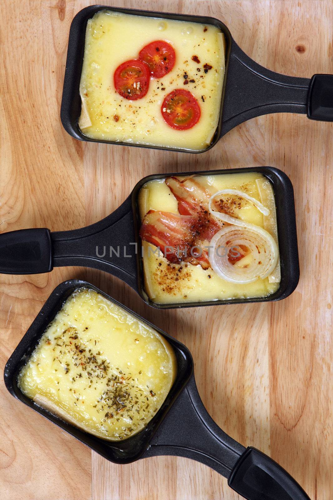 Photo of a three Raclette trays with melted cheese, herbs, cherry tomato, bacon and onion.