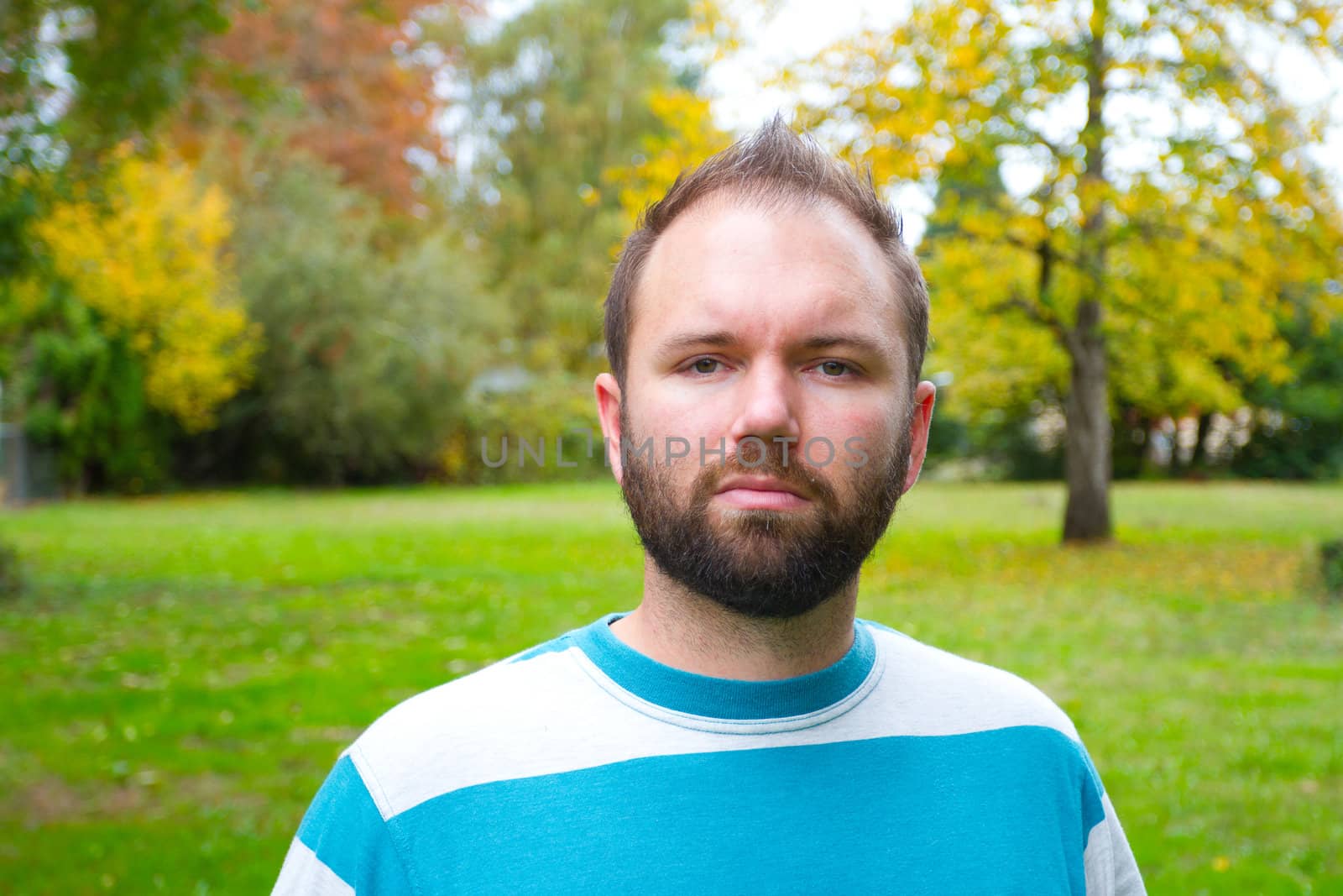 A man with a full beard looks directly at the camera while outdoors in a park setting.