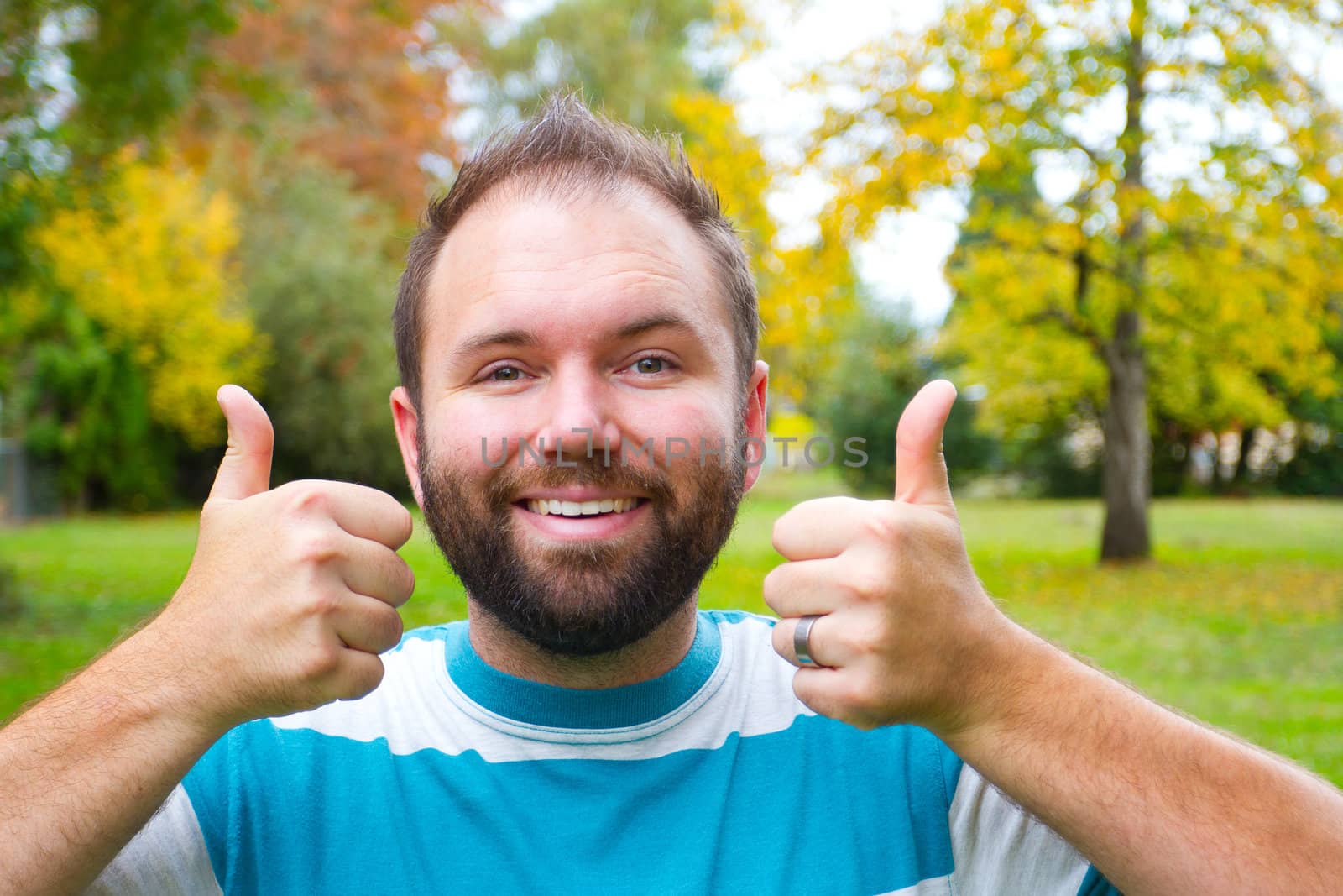 A man with a full beard gives the camera a double thumgs up with both hands while outdoors in a park setting.