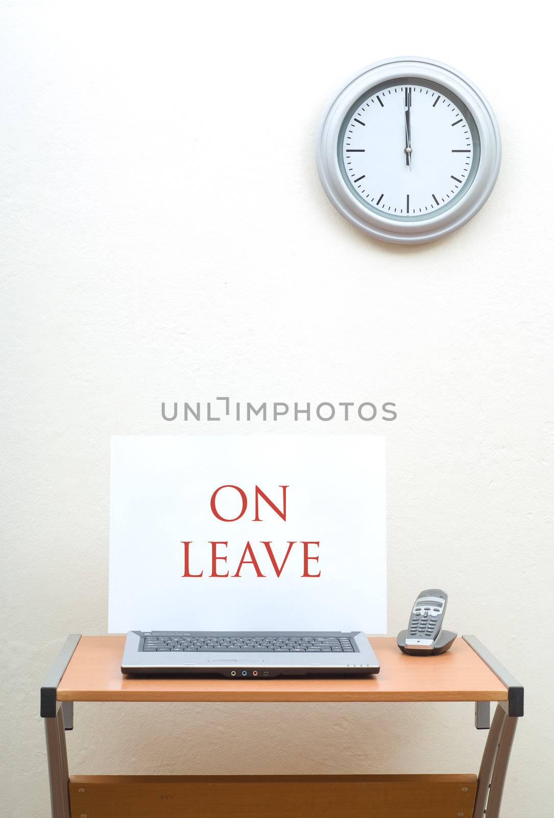 Office desk with on leave sign on open laptop with phone, clock on wall