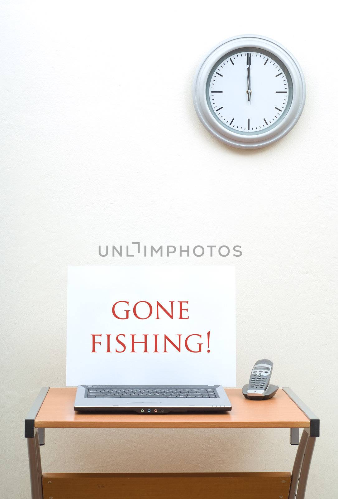Office desk with gone fishing sign on open laptop next to portable phone, clock on wall