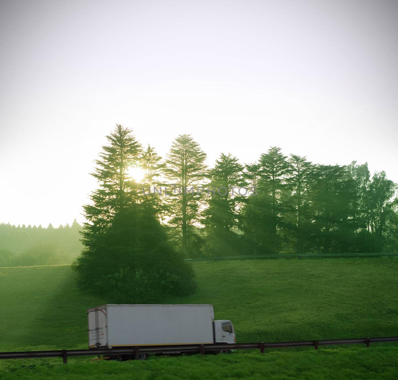 Sun and forest trees shine down on white truck delivery with motion blur on highway road