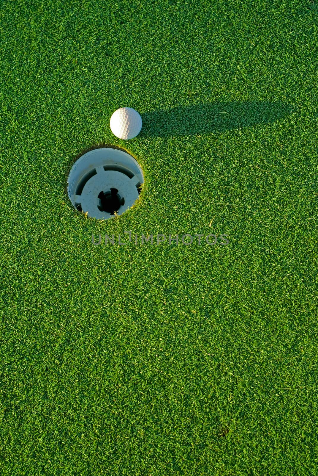 White golf ball on putting green next to hole with long shadow - from top side down.