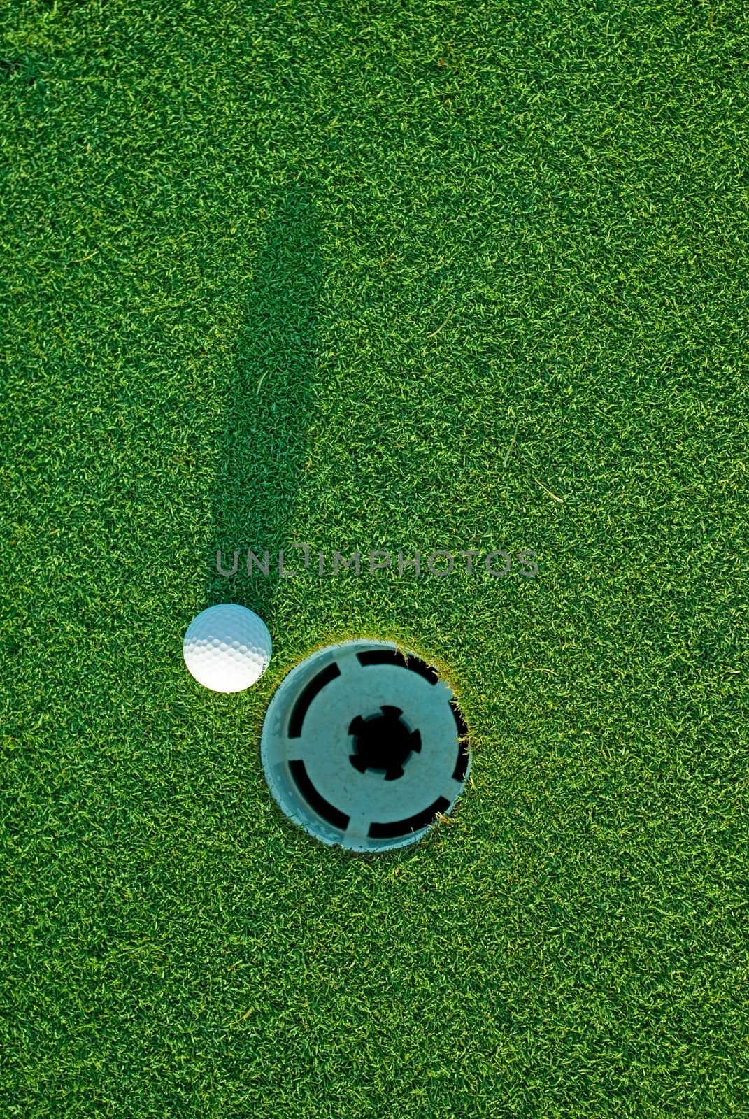 White golf ball on putting green next to hole with long shadow - from top down.
