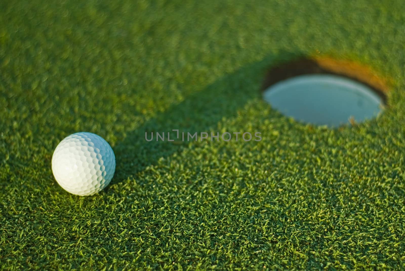 White golf ball on putting green next to hole with long shadow and selective focus on ball