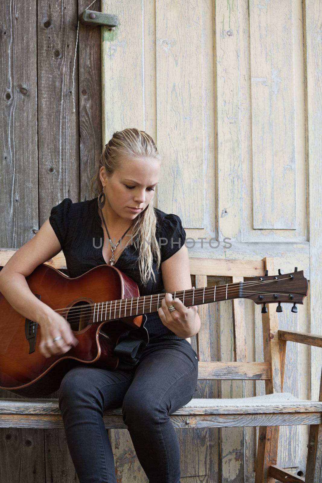 Photo of a beautiful blond female playing her acoustic guitar on an old wooden bench. 