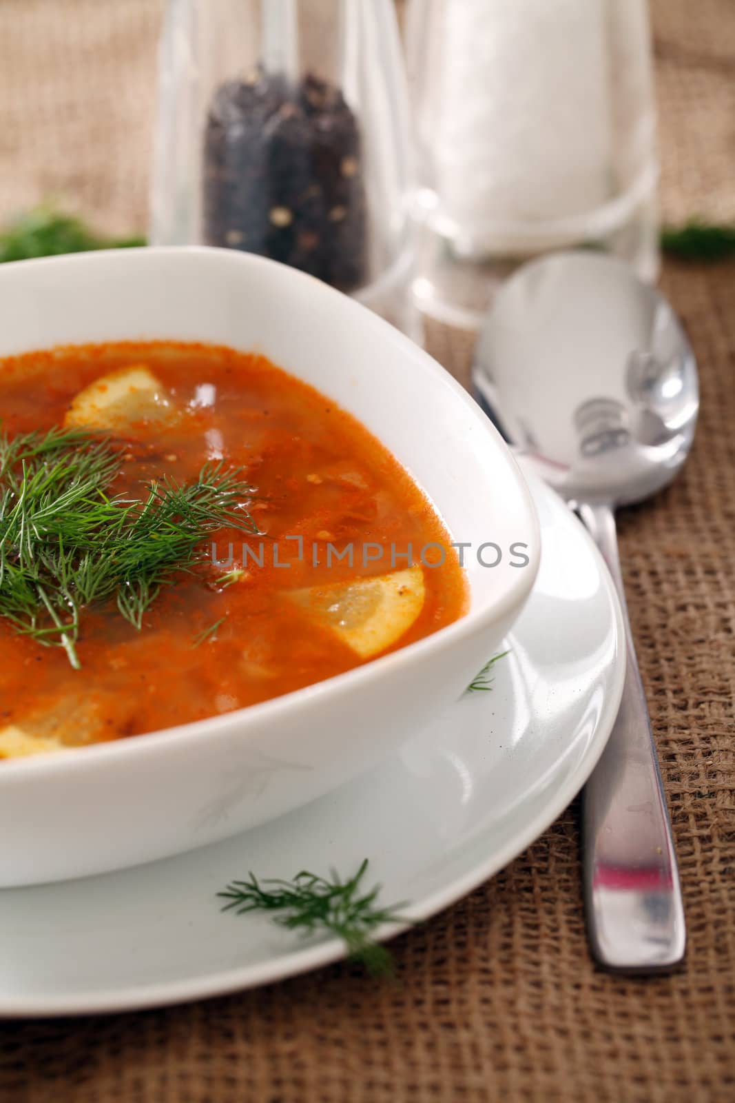 Image of bowl of hot red soup served with the salt, pepper and spoon on a beige tablecloth