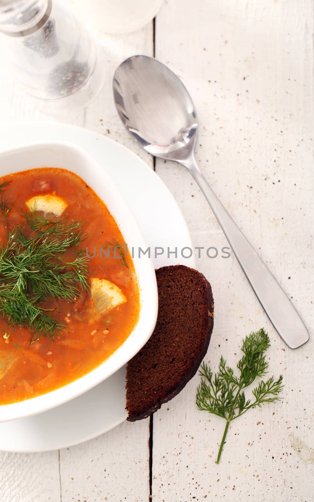 Image of bowl of hot red soup served with bead, pepper, salt and spoon on white wooden table