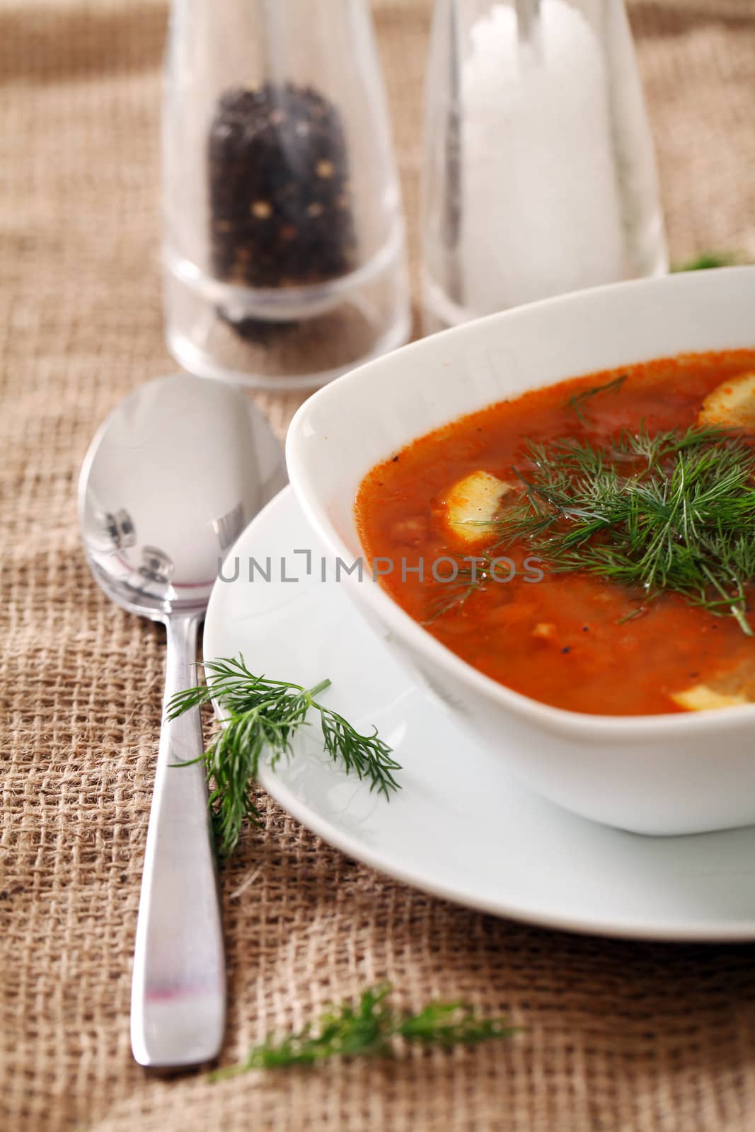 Image of bowl of hot red soup served with the salt, pepper and spoon on a beige tablecloth
