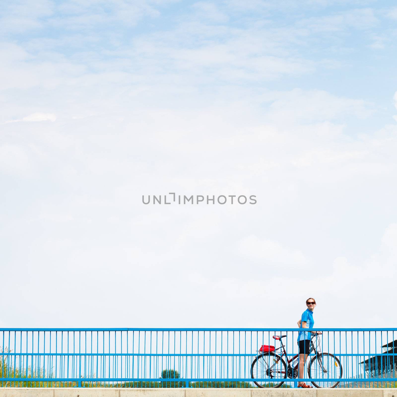 Background for poster or advertisment pertaining to cycling/sport/outdoor activities - female cyclist during a halt on a bridge against blue sky (color toned image)