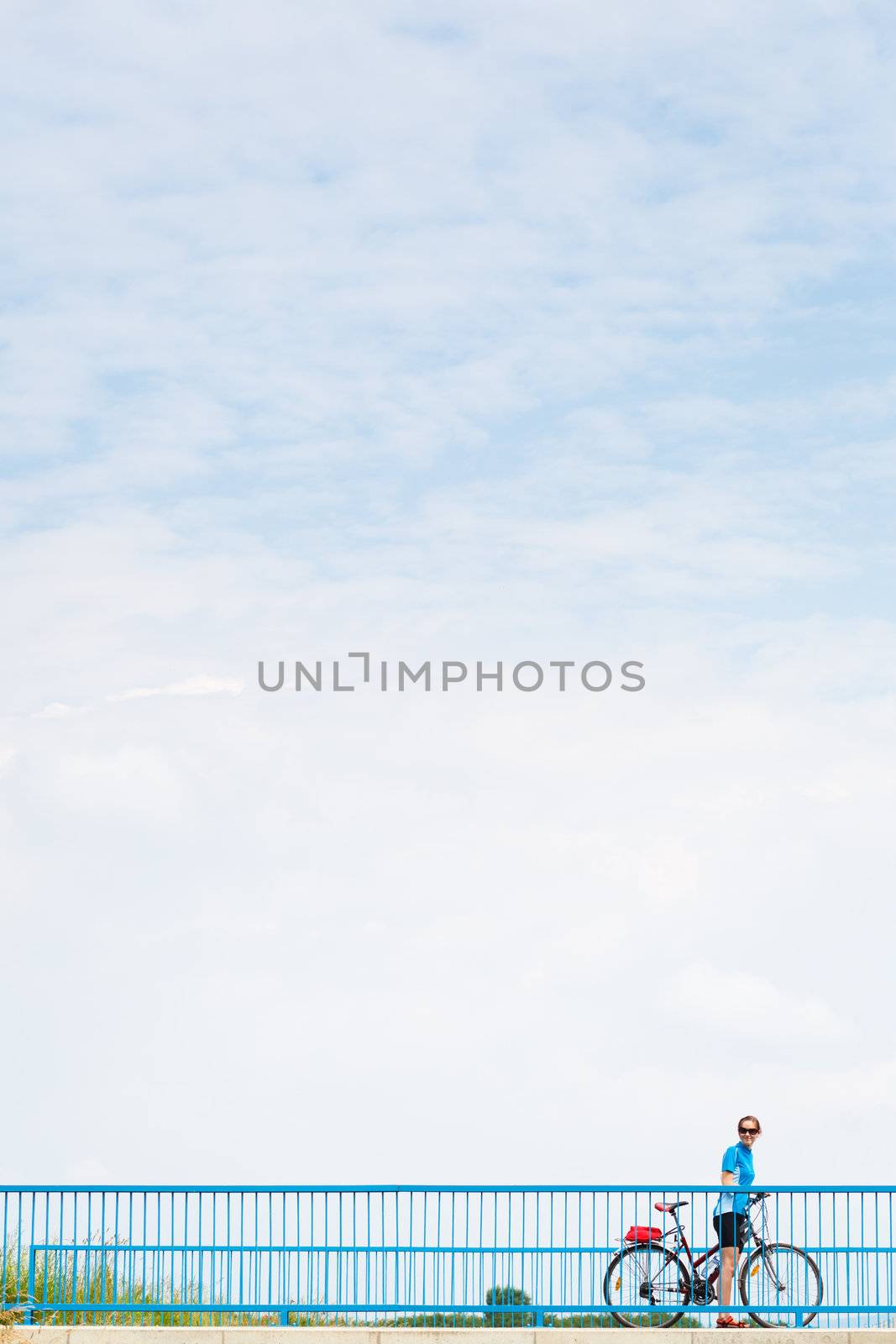 Background for poster or advertisment pertaining to cycling/sport/outdoor activities - female cyclist during a halt on a bridge against blue sky (color toned image)