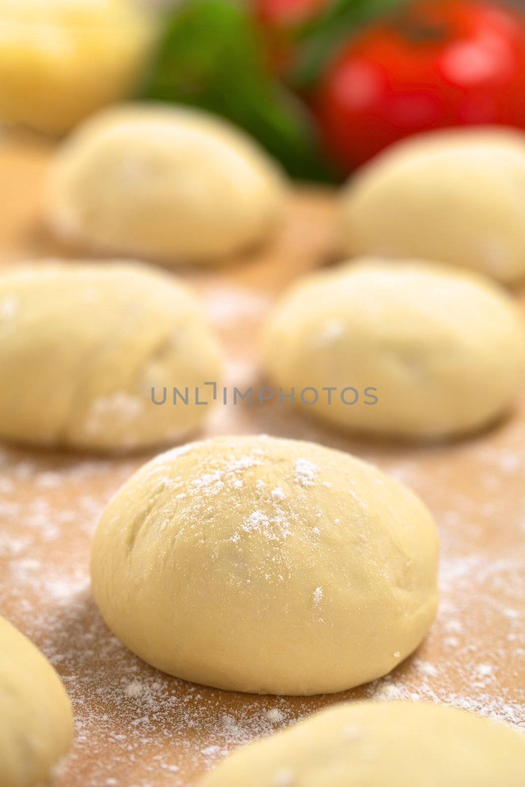 Small balls of fresh homemade pizza dough on floured wooden board with pizza ingredients (tomato, basil, grated cheese) in the back (Selective Focus, Focus one third into the first pizza dough) 