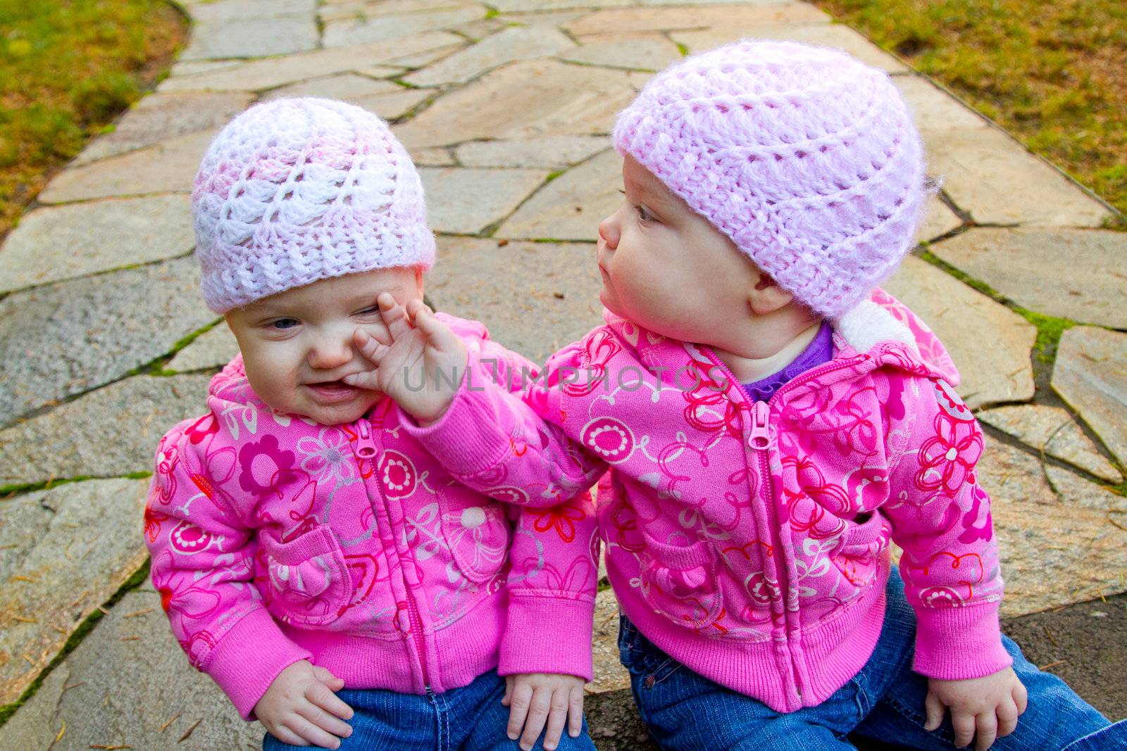 Two twin baby girls sit on a stone walkway wearing pink sweatshirts and beanies.