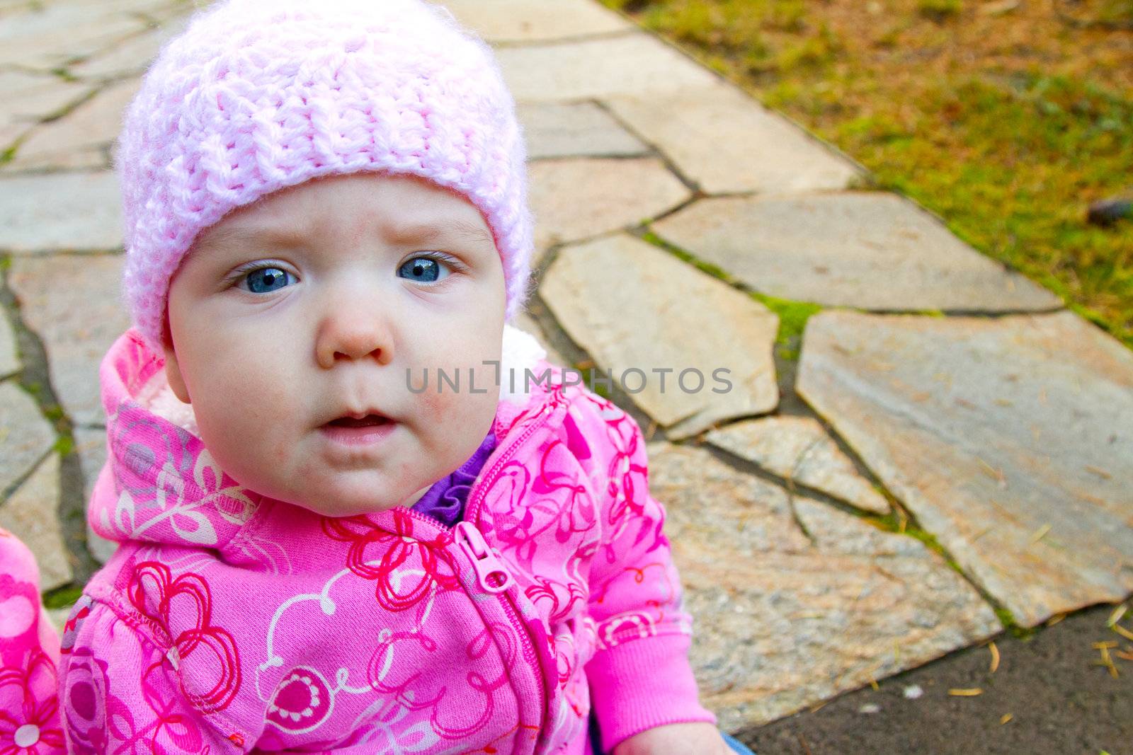 A young infant girl wearing a pink sweatshirt and a knitted cap sits on a stone walkway for a portrait.