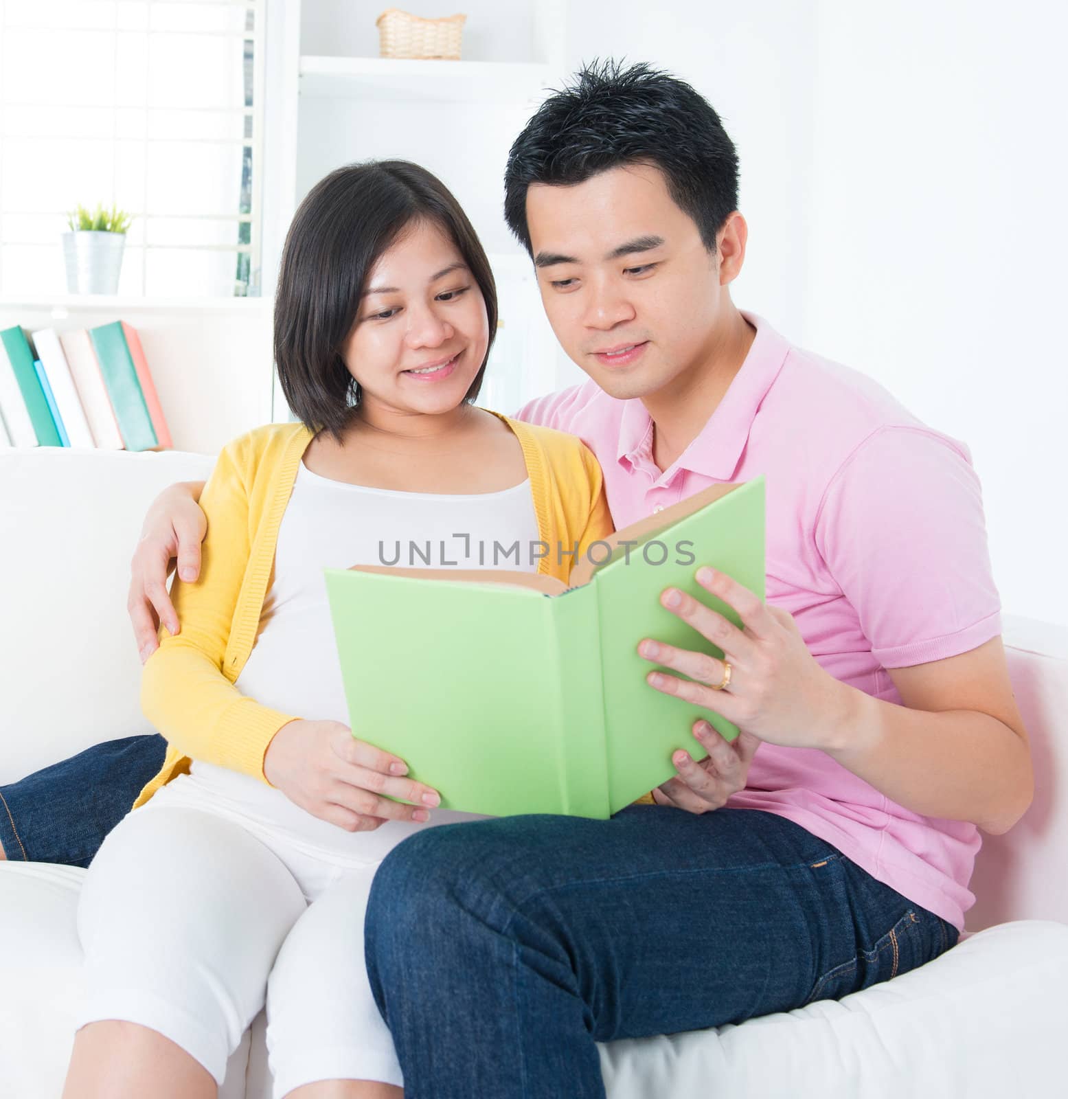 Asian couple reading book together, sitting on sofa