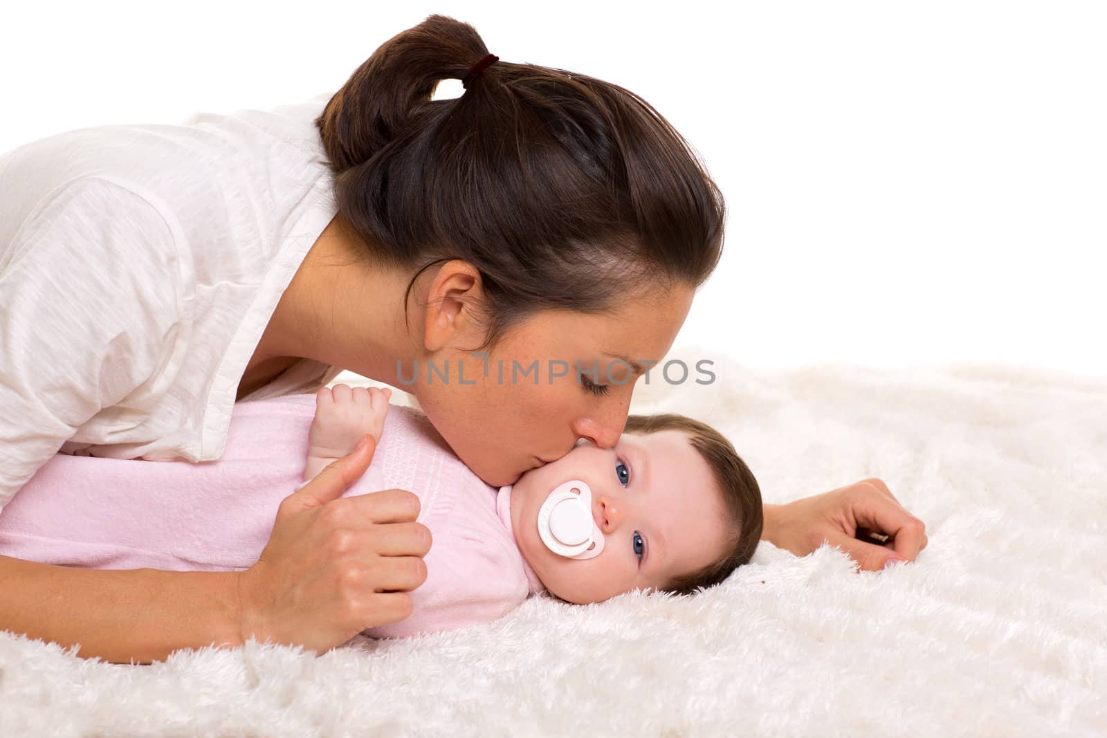 Baby girl and mother kissing her lying happy on white fur blanket