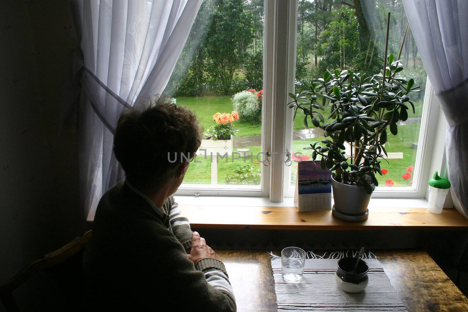 An old, lonely lady, sitting next to her kitchen table, looks out of her window. It is raining outside. Horizontal picture