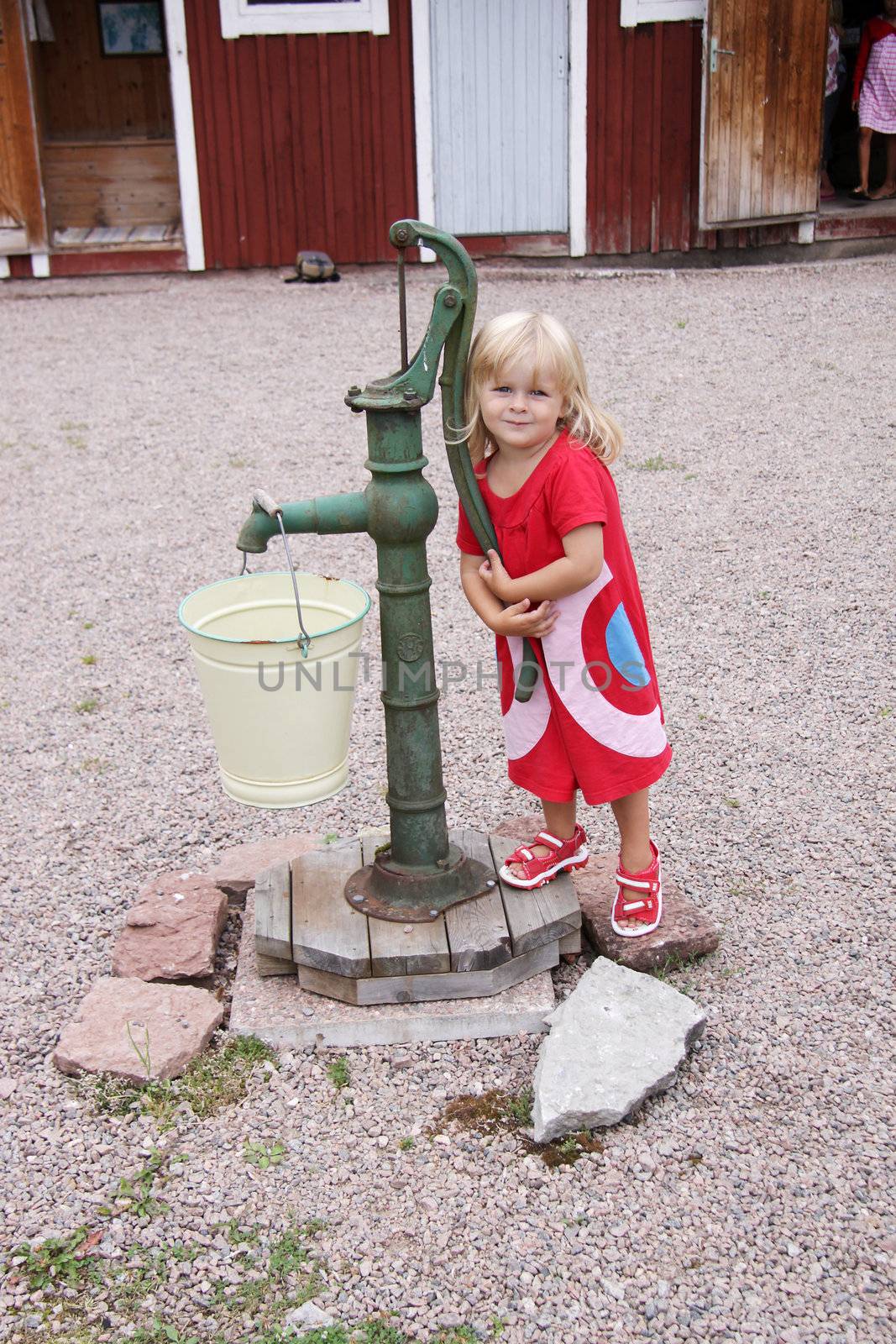 A cute blonde little girl standing next to a waterpump at an outdoor museum