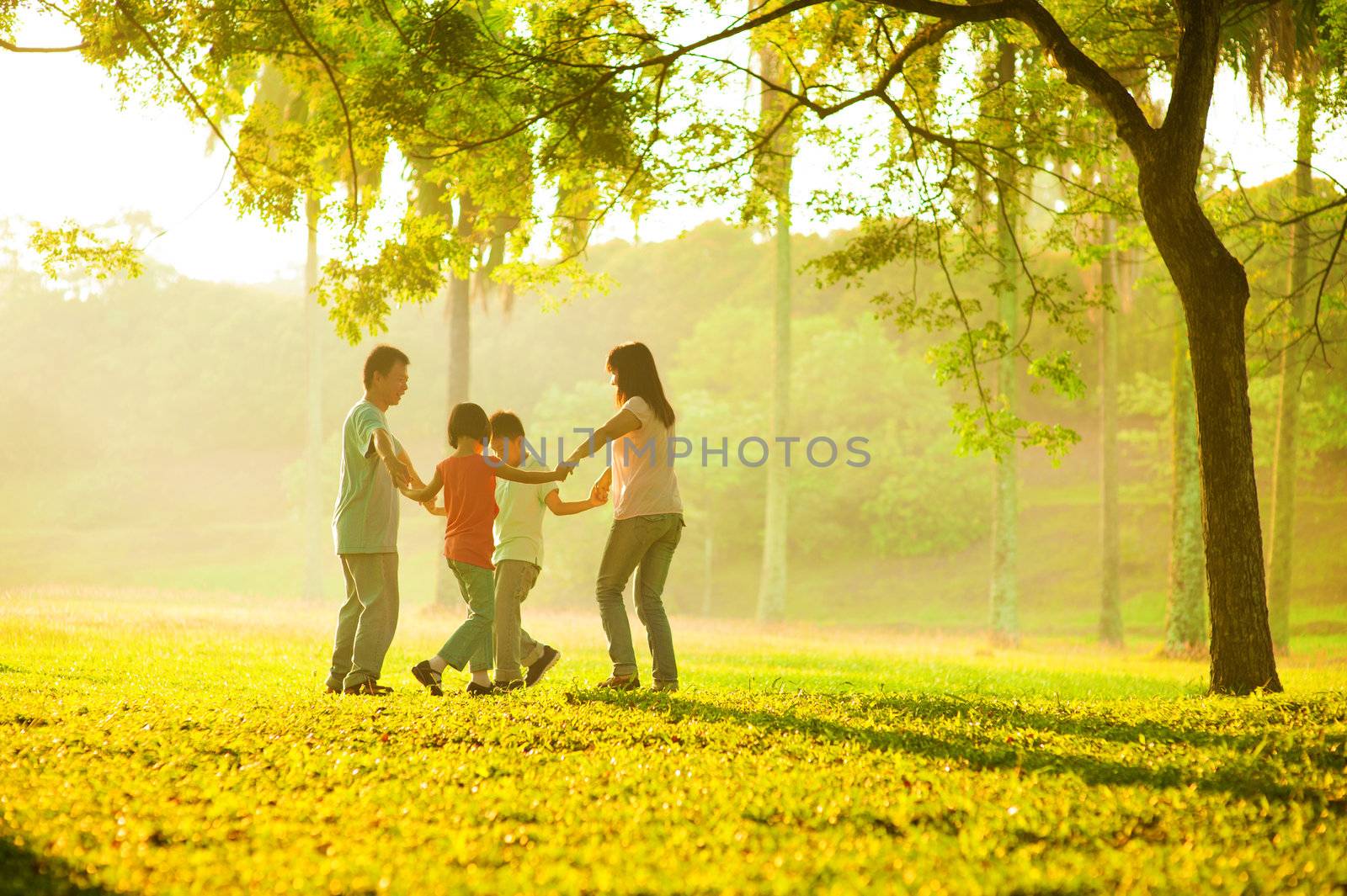 Happy asian family playing on the field during sunrise. People running in circle under the tree at outdoor green park.