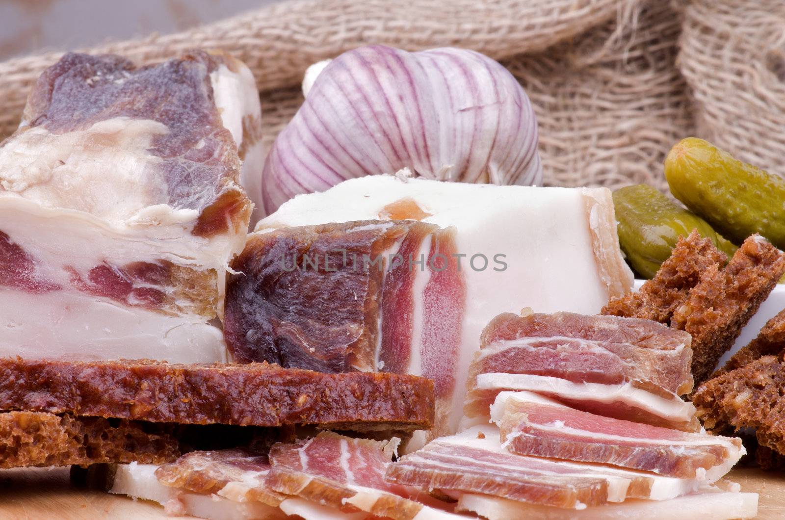 Arrangement of Raw Smoked Homemade Bacon with Brown Bread, Garlic and and Marinated Gherkins closeup on Sacking background