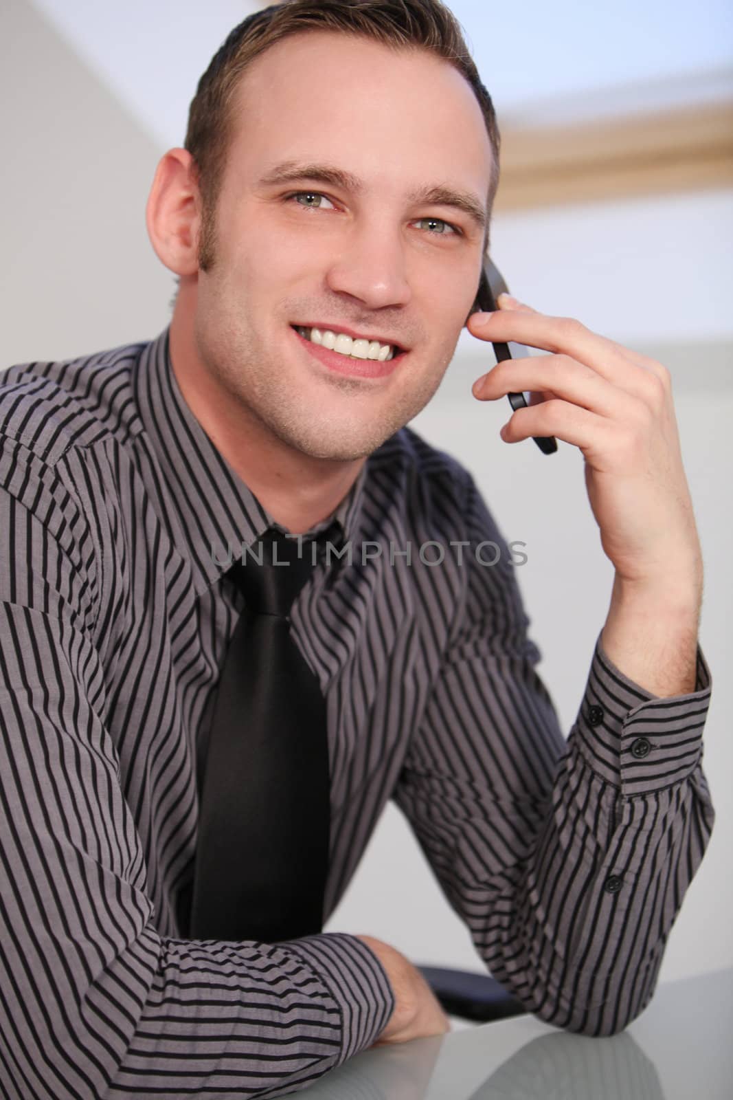 Close-up of a young business man smiling at the camera and talking on a smartphone. Smiling young man in a stylish shirt and tie sitting at a table using a smartphone or mobile phone