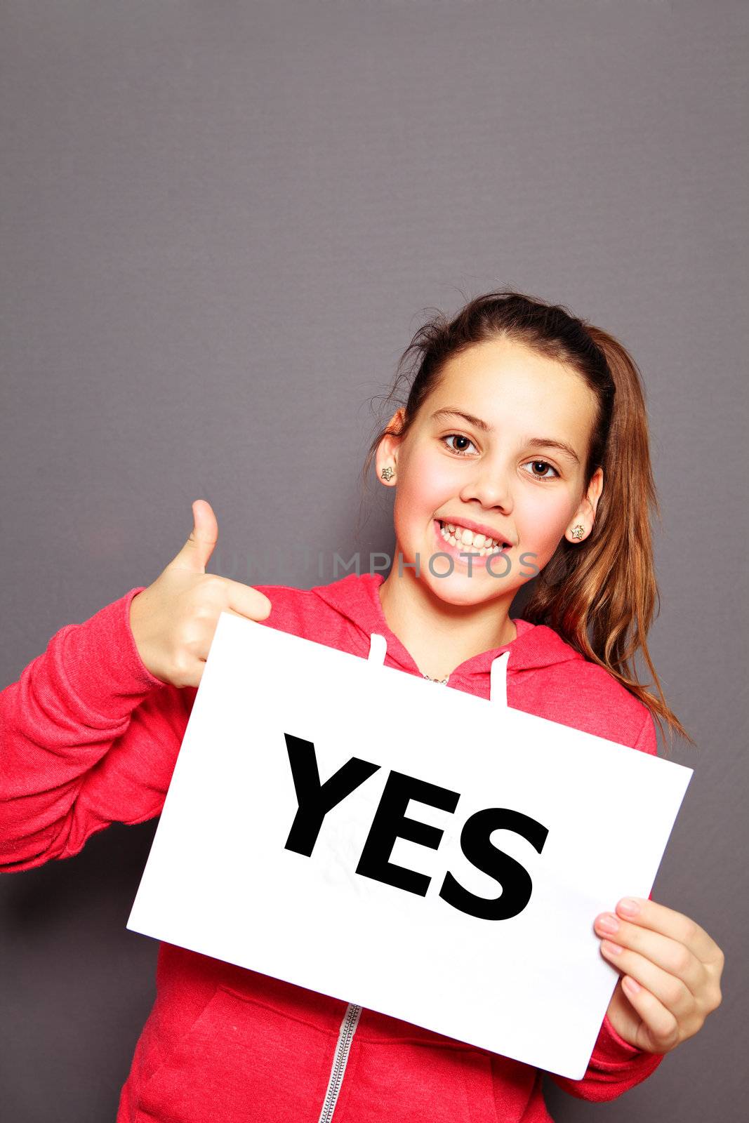 Beautiful enthusiastic happy little girl with a typewritten YES sign giving a thumbs up gesture of approval and agreement, studio upper body portrait on a grey background with copyspace