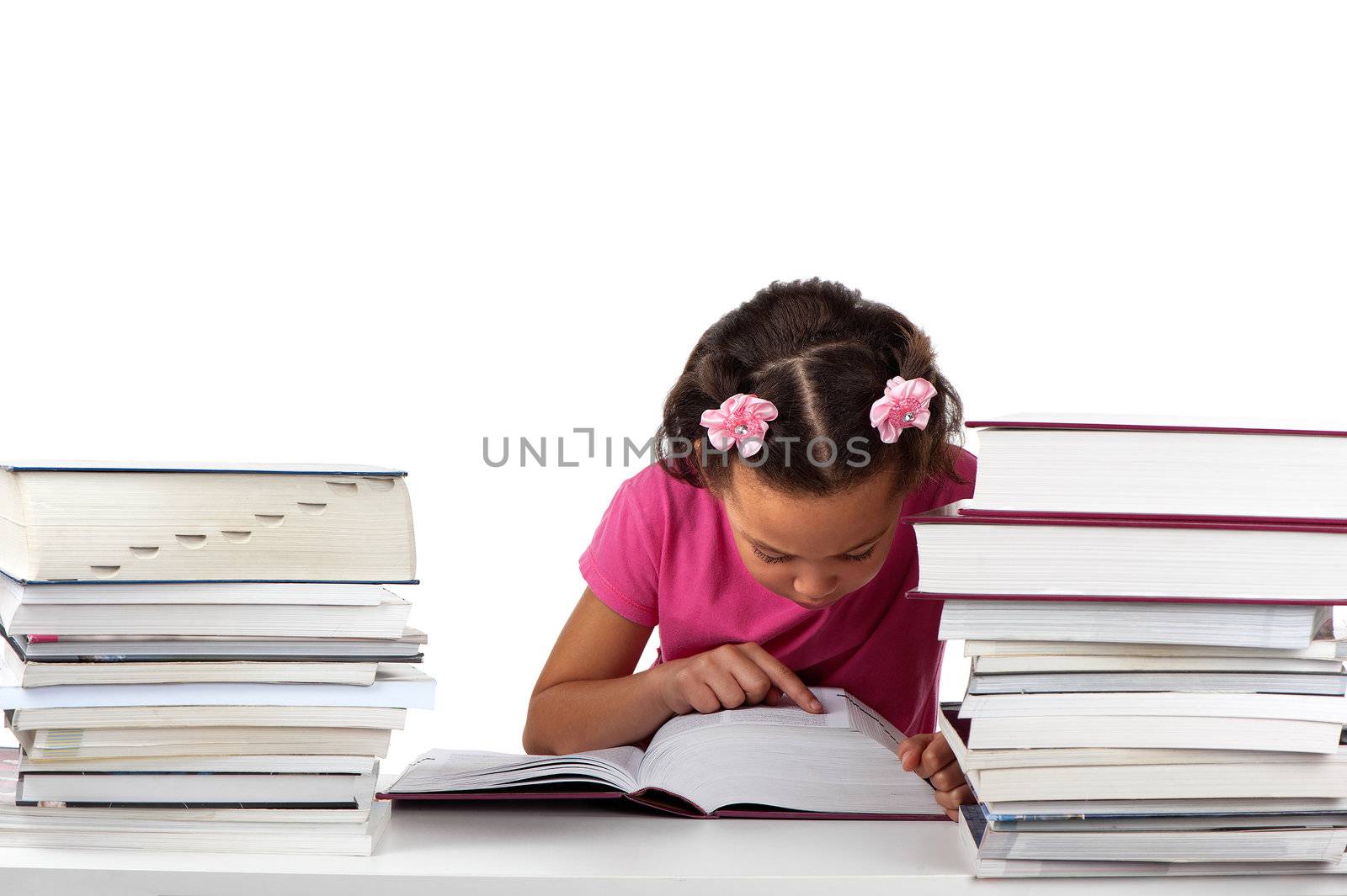 A young girl stays focused whilst reading a huge book in the midst of other reading material.