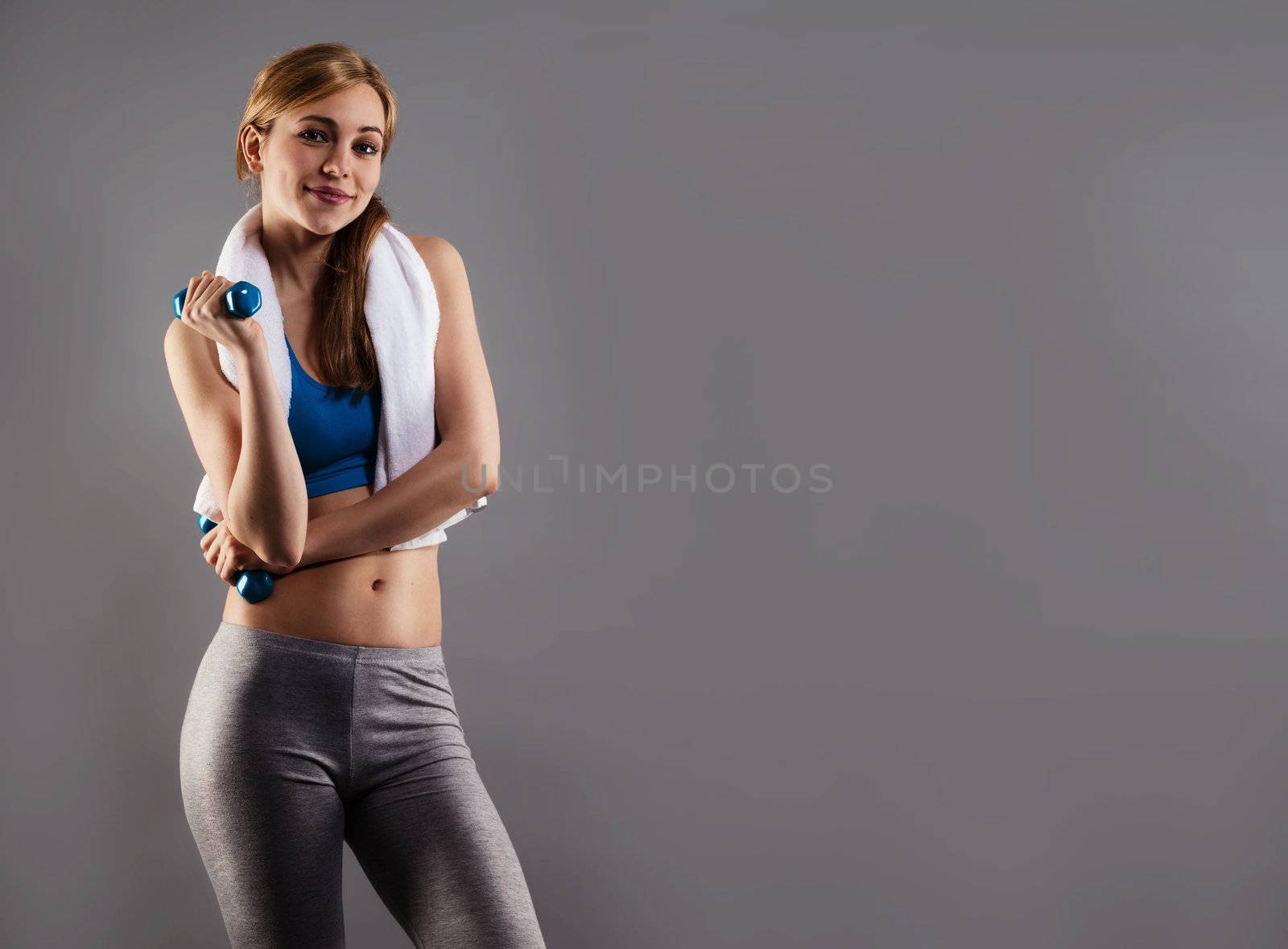 smiling fitness woman with a white towel and blue dumbbells on gray background