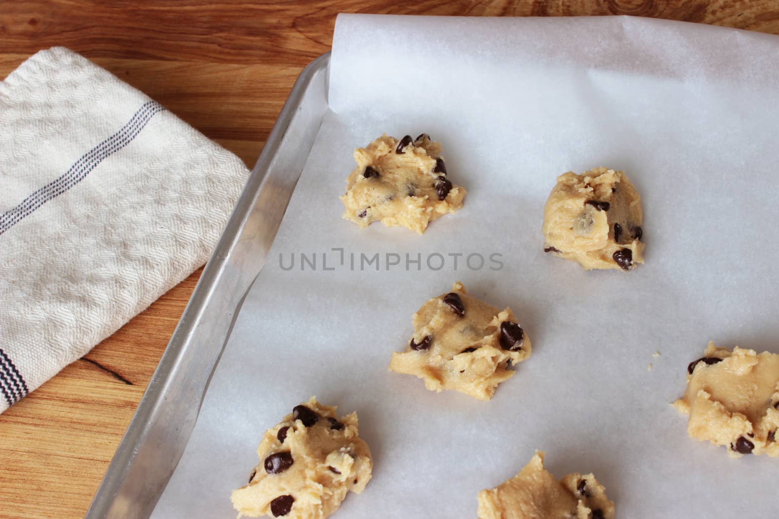A close-up image of a cookie sheet lined with parchment paper, with chocolate chip cookie dough shaped into cookies, on a wooden counter with a white dish towel.