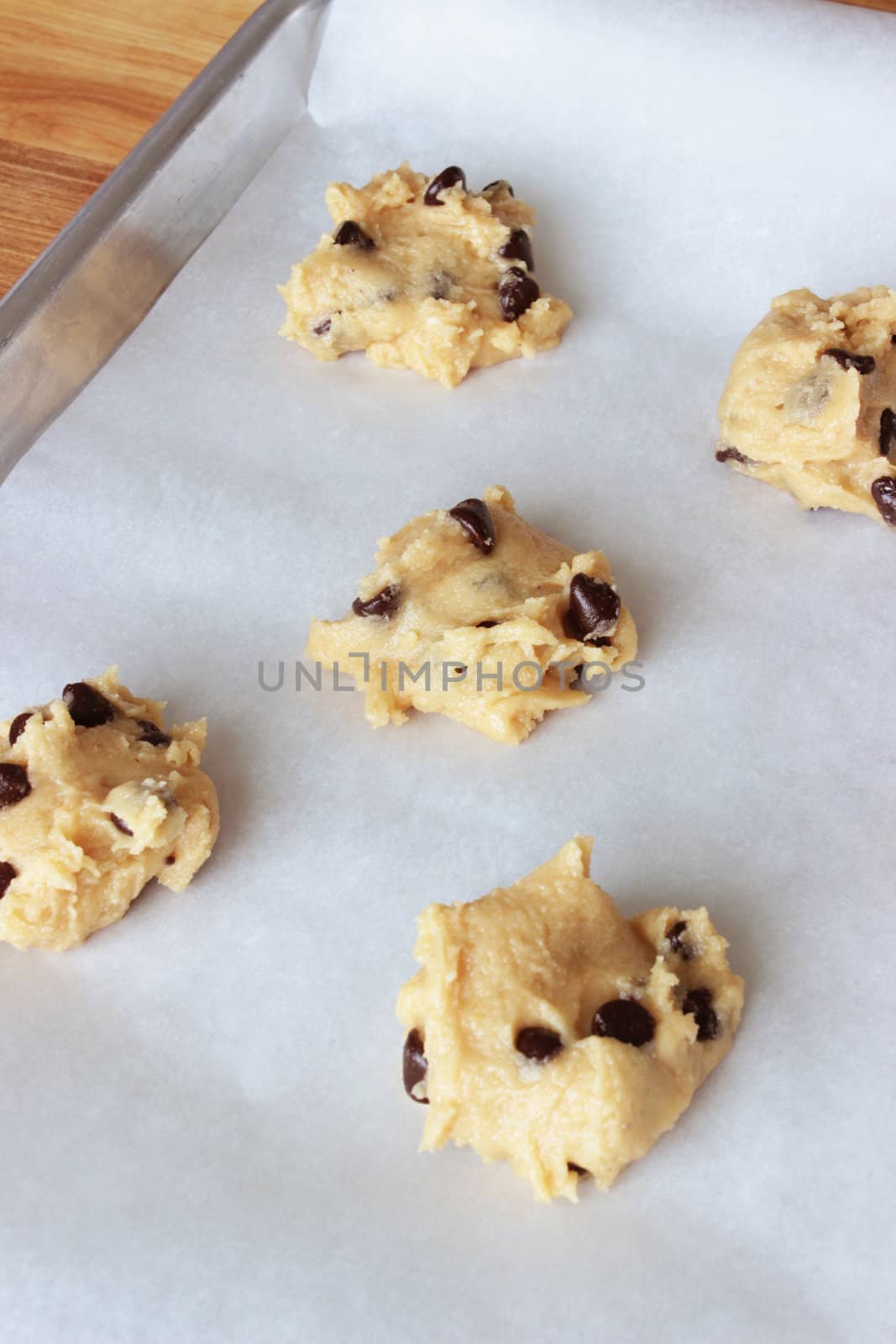 A close-up image of a cookie sheet lined with parchment paper, with chocolate chip cookie dough shaped into cookies, on a wooden counter.