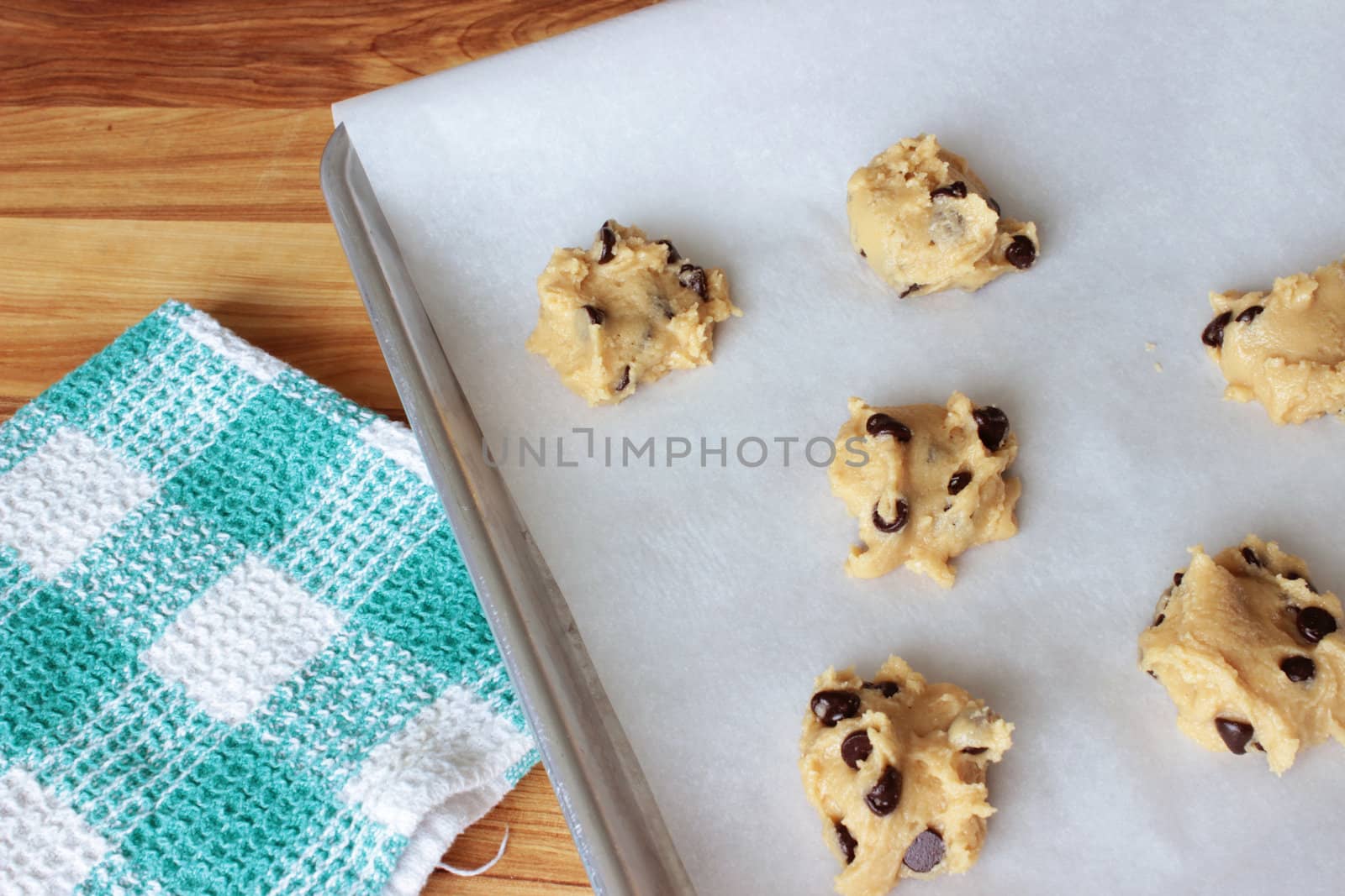 A close-up image of a cookie sheet lined with parchment paper, with chocolate chip cookie dough shaped into cookies, on a wooden counter with a green and white dish towel.
