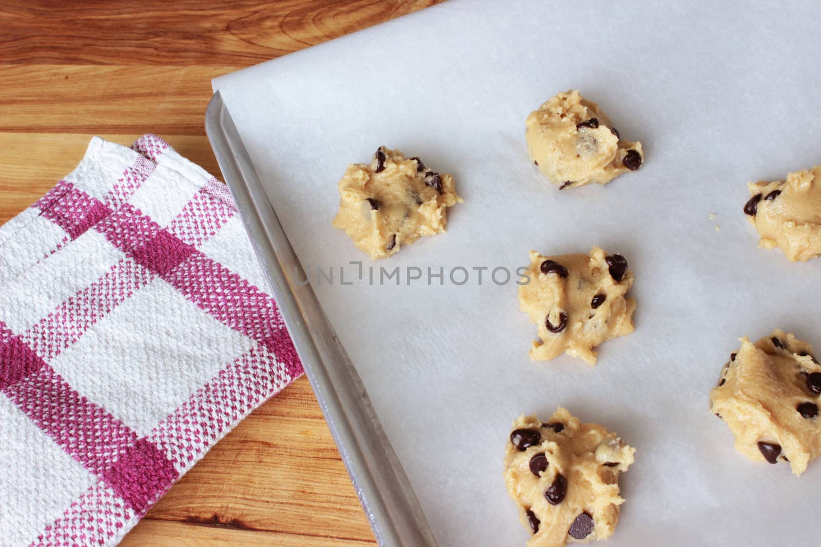 A close-up image of a cookie sheet lined with parchment paper, with chocolate chip cookie dough shaped into cookies, on a wooden counter with a red and white dish towel.