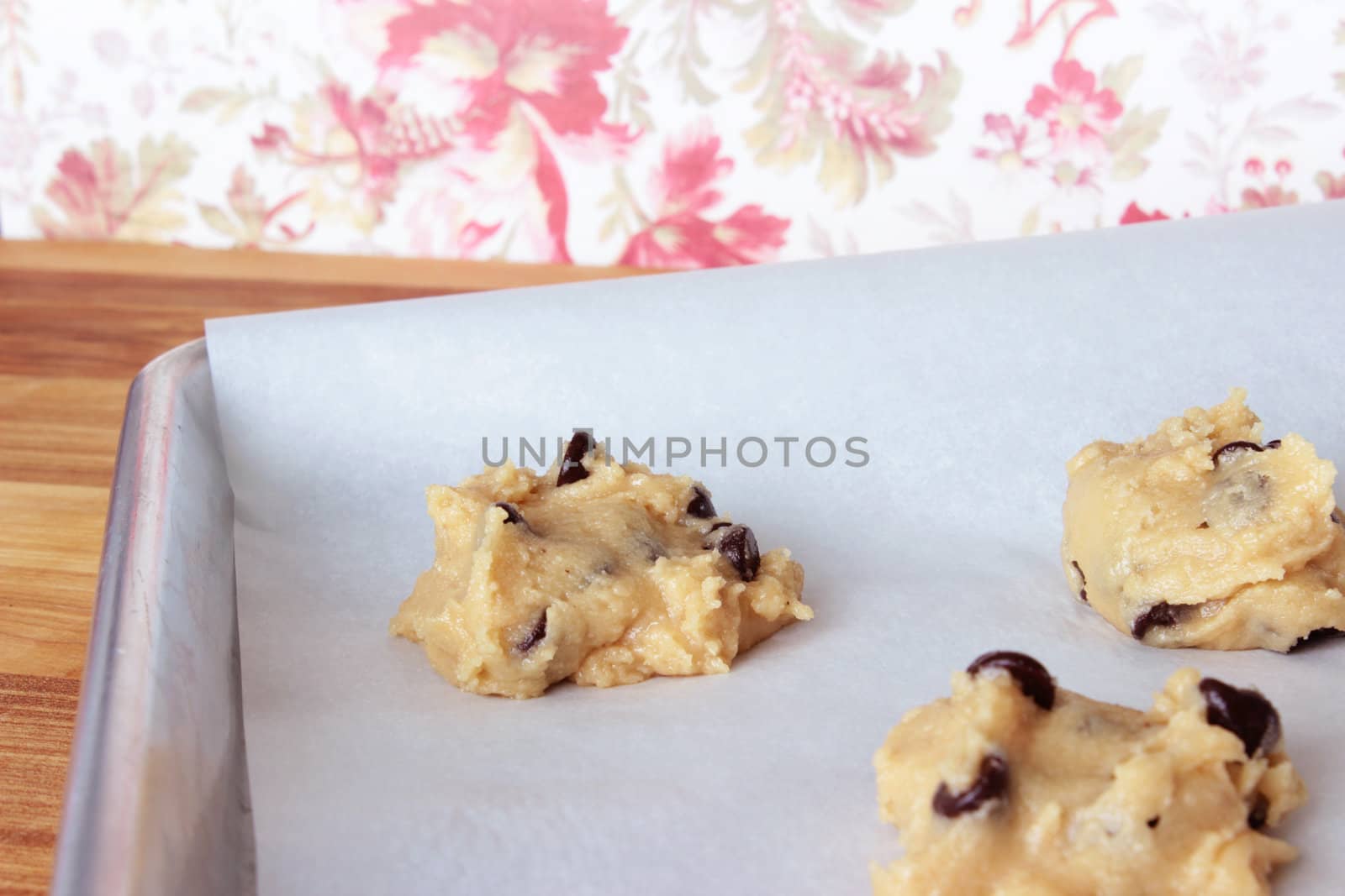 A close-up image of a cookie sheet lined with parchment paper, with chocolate chip cookie dough shaped into cookies, on a wooden counter with a red and white vintage flowered wallpaper background.