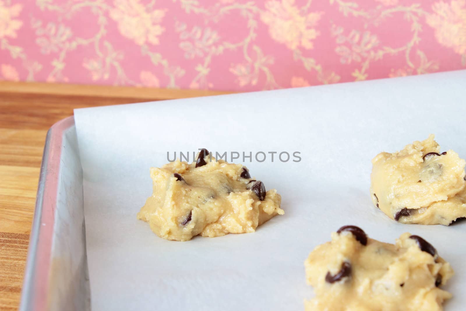 A close-up image of a cookie sheet lined with parchment paper, with chocolate chip cookie dough shaped into cookies, on a wooden counter with a dark pink vintage flowered wallpaper background.