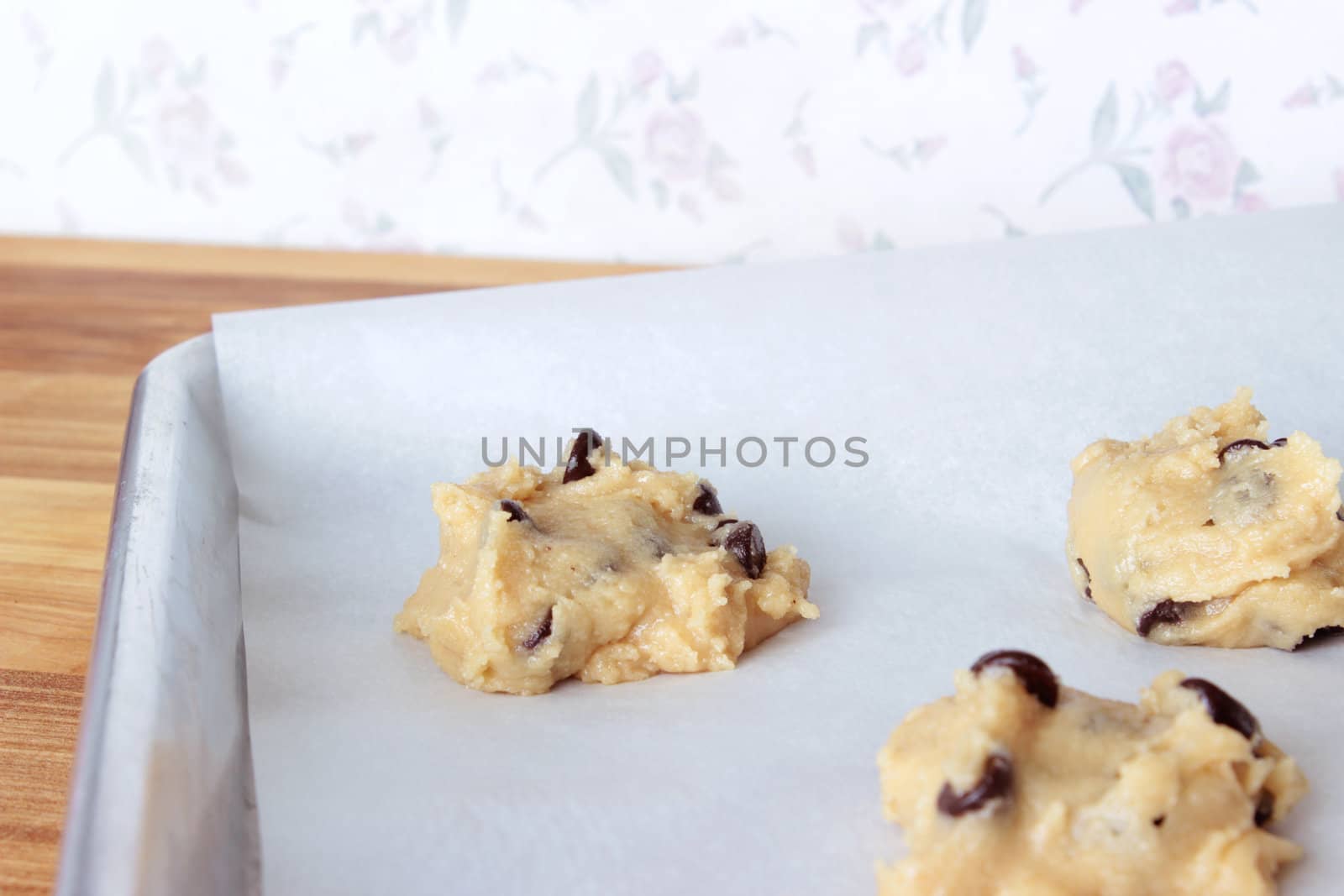 A close-up image of a cookie sheet lined with parchment paper, with chocolate chip cookie dough shaped into cookies, on a wooden counter with a vintage white wallpaper background with pink flowers.