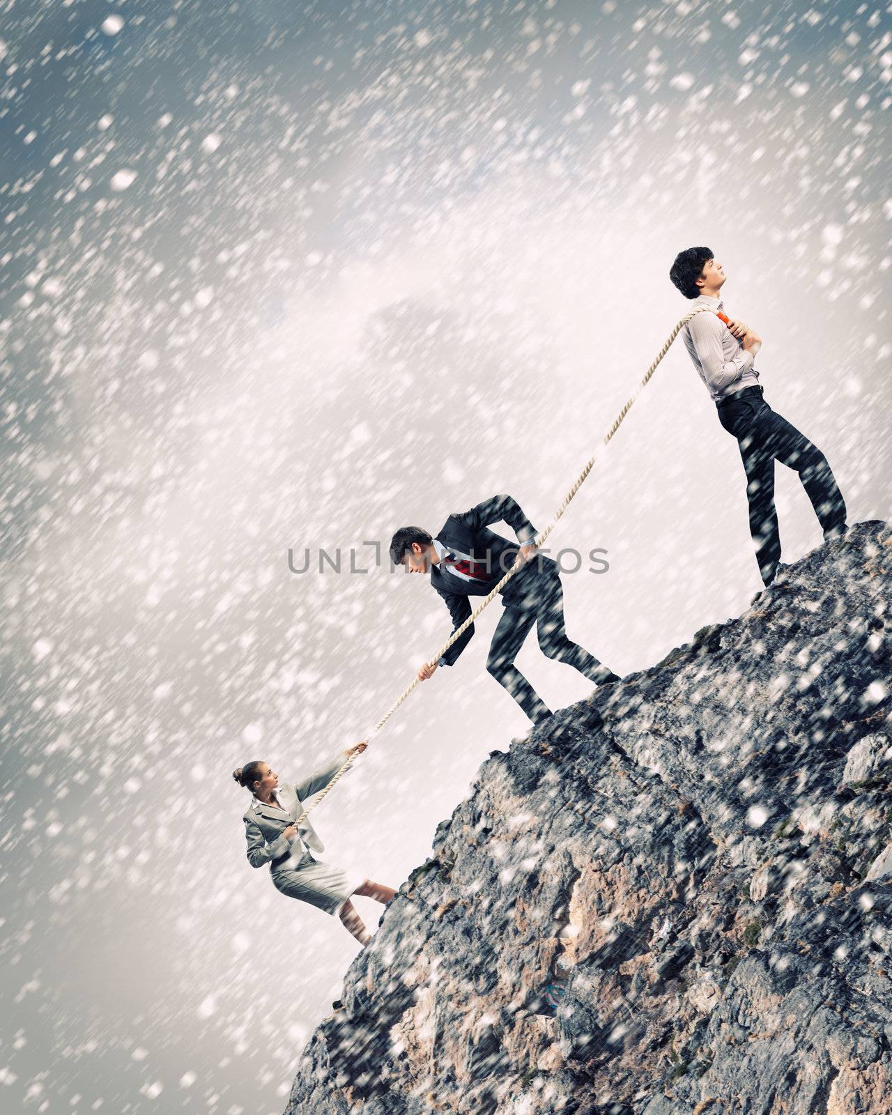 Image of three businesspeople pulling rope atop of mountain under falling snow