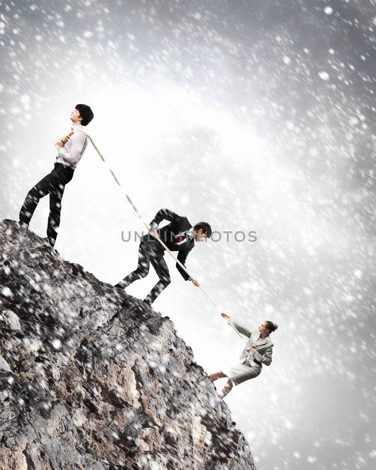 Image of three businesspeople pulling rope atop of mountain under falling snow