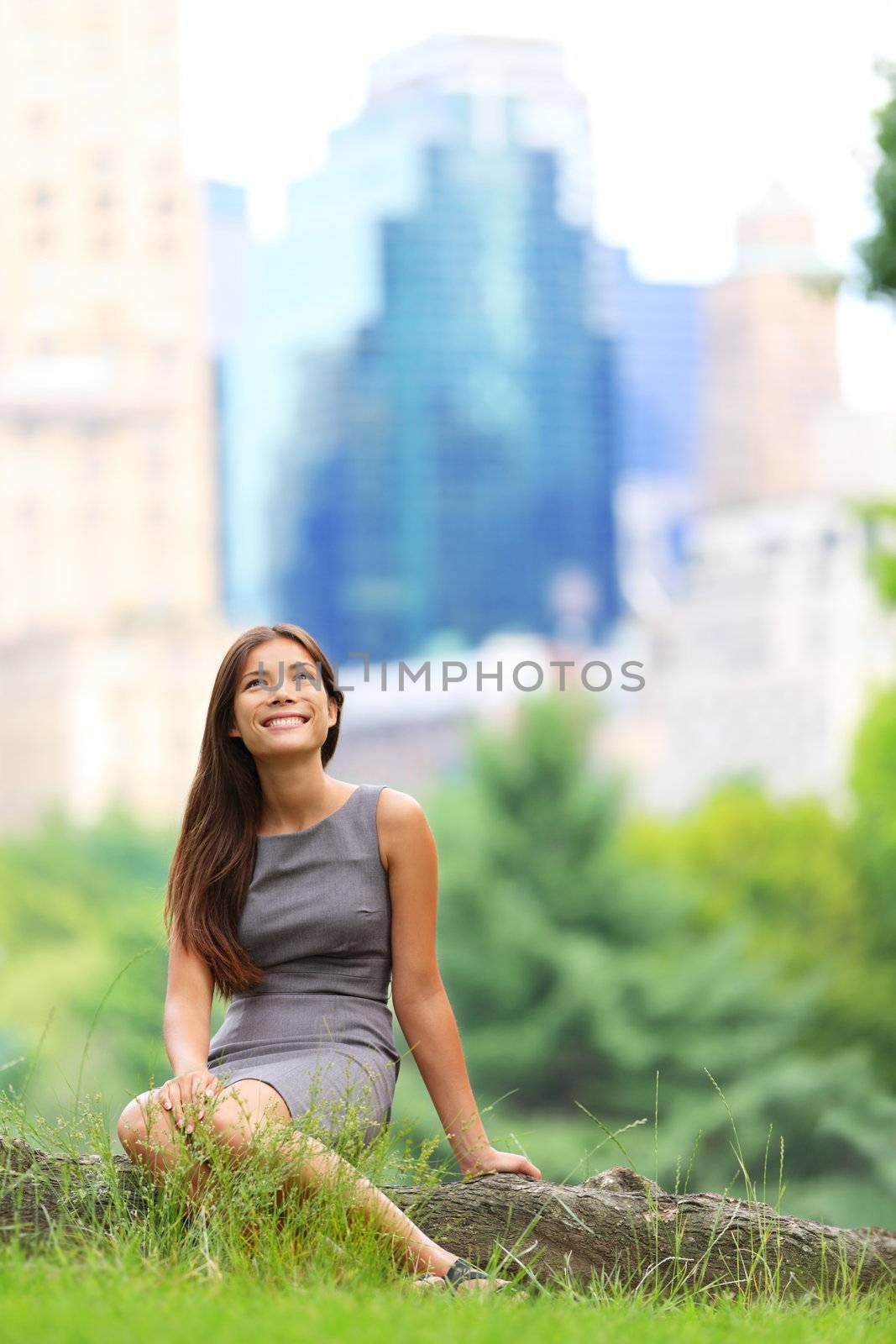 Young Asian Business woman in New York Central Park. Businesswoman in New York City, Manhattan, Central Park sitting looking up at copy space with skyscraper buildings from New York skyline in background. Young female professional. Mixed Caucasian / Asian race.