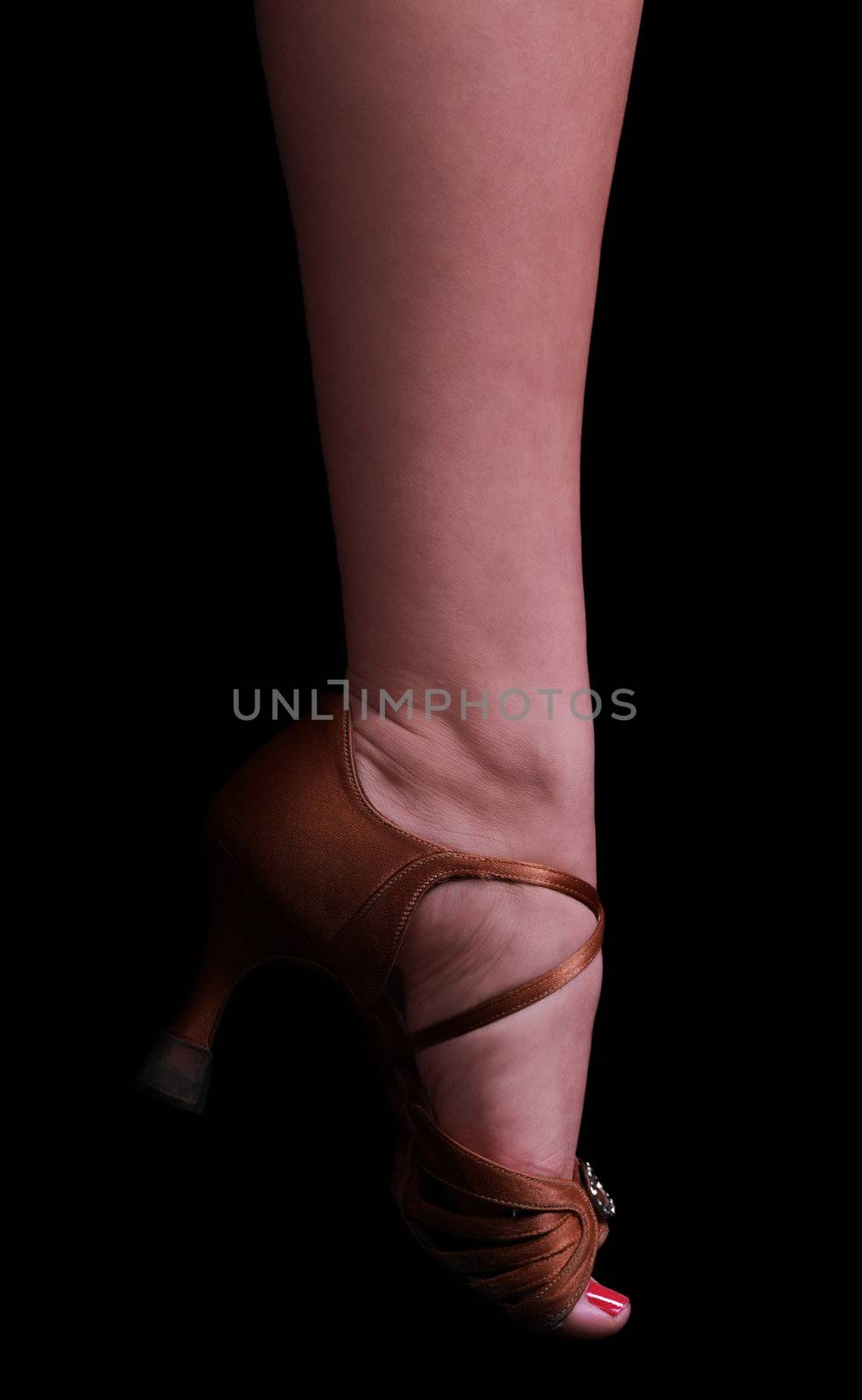 The foot of a female dancer in a high heel dancing sandal isolated aginst a black background.