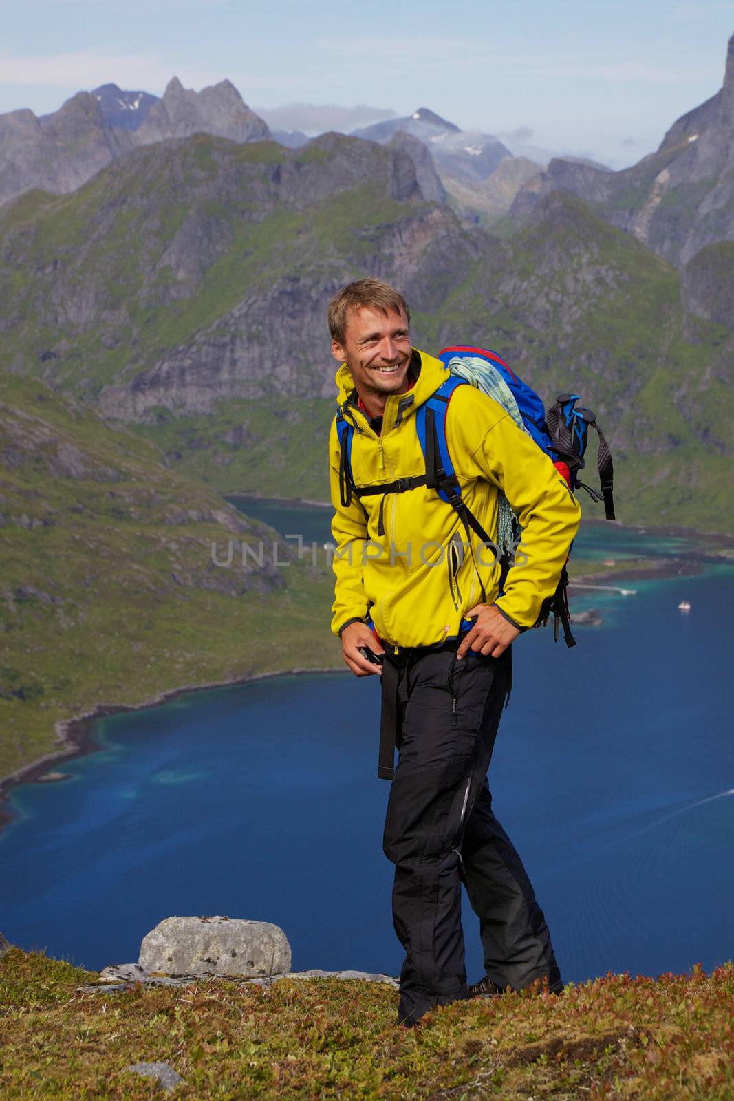 Young smiling active man with backpack hiking on Lofoten islands in Norway on sunny day high above fjord