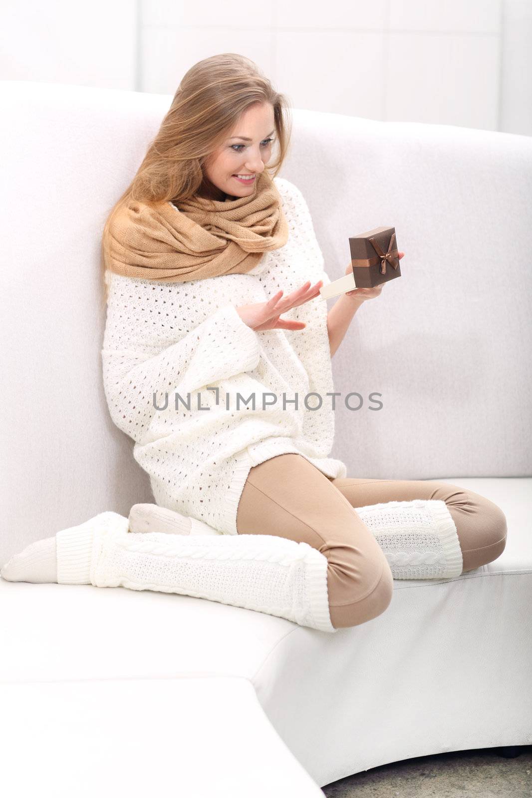 Long-haired young blonde woman sitting on a sofa. He holds a box with a present. She is surprised and very happy.